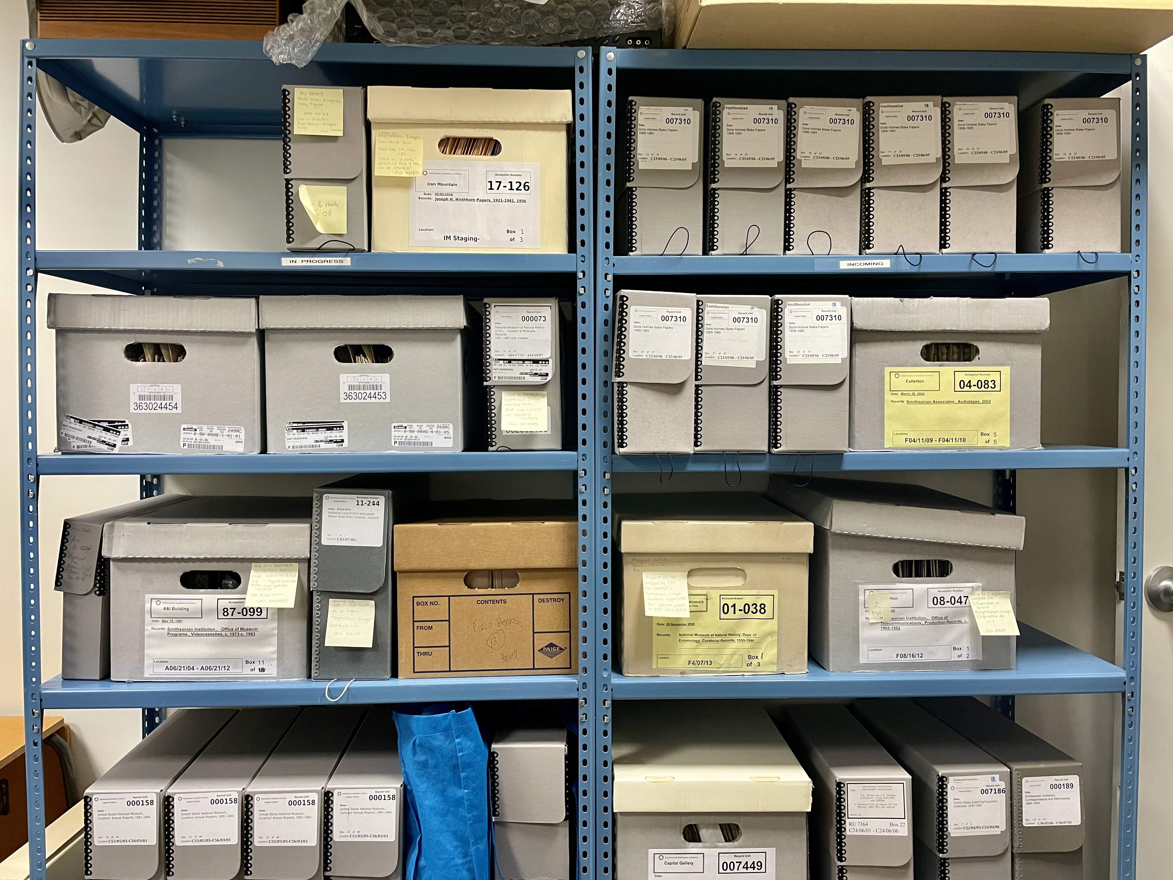 To locate collections that are not in their regular storage location, staff have needed to check other storage locations, including shelves in the imaging labs. A pair of blue metal shelves filled with an assortment of archival boxes of varying sizes. Self-adhesive notes on the exteriors indicate project progress.