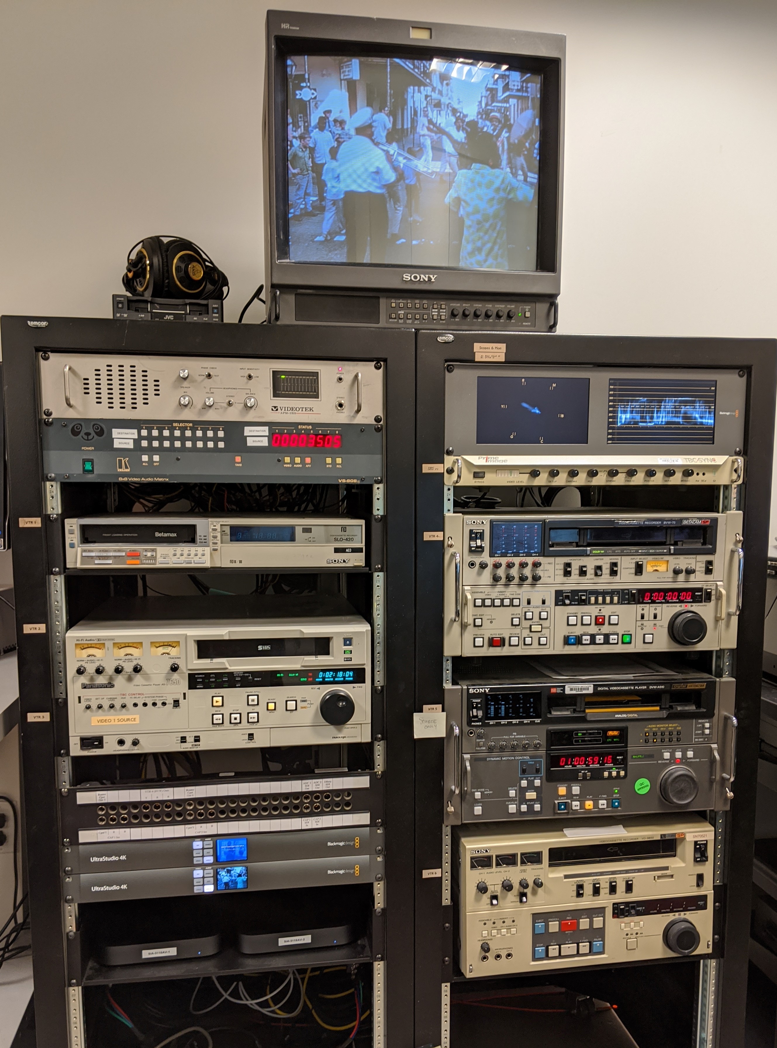 This is our new video digitization rack. The analog-to-digital converters and Mac Minis have replaced the previous equipment. Courtesy of Kira Sobers. A rack of assorted legacy video decks. A monitor sitting on top of the rack shows a screenshot of a video of a parade in the street.