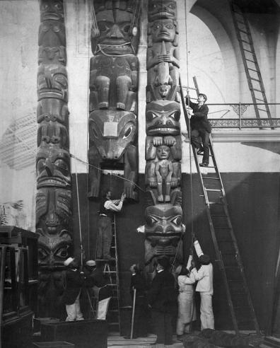 Curators look on as three totem poles are installed in the Northwest Range near the Rotunda of the United States National Museum, now the Arts and Industries Building, by several laborers. Record Unit 95, Smithsonian Institution Archives, Neg. no. MNH-5792.