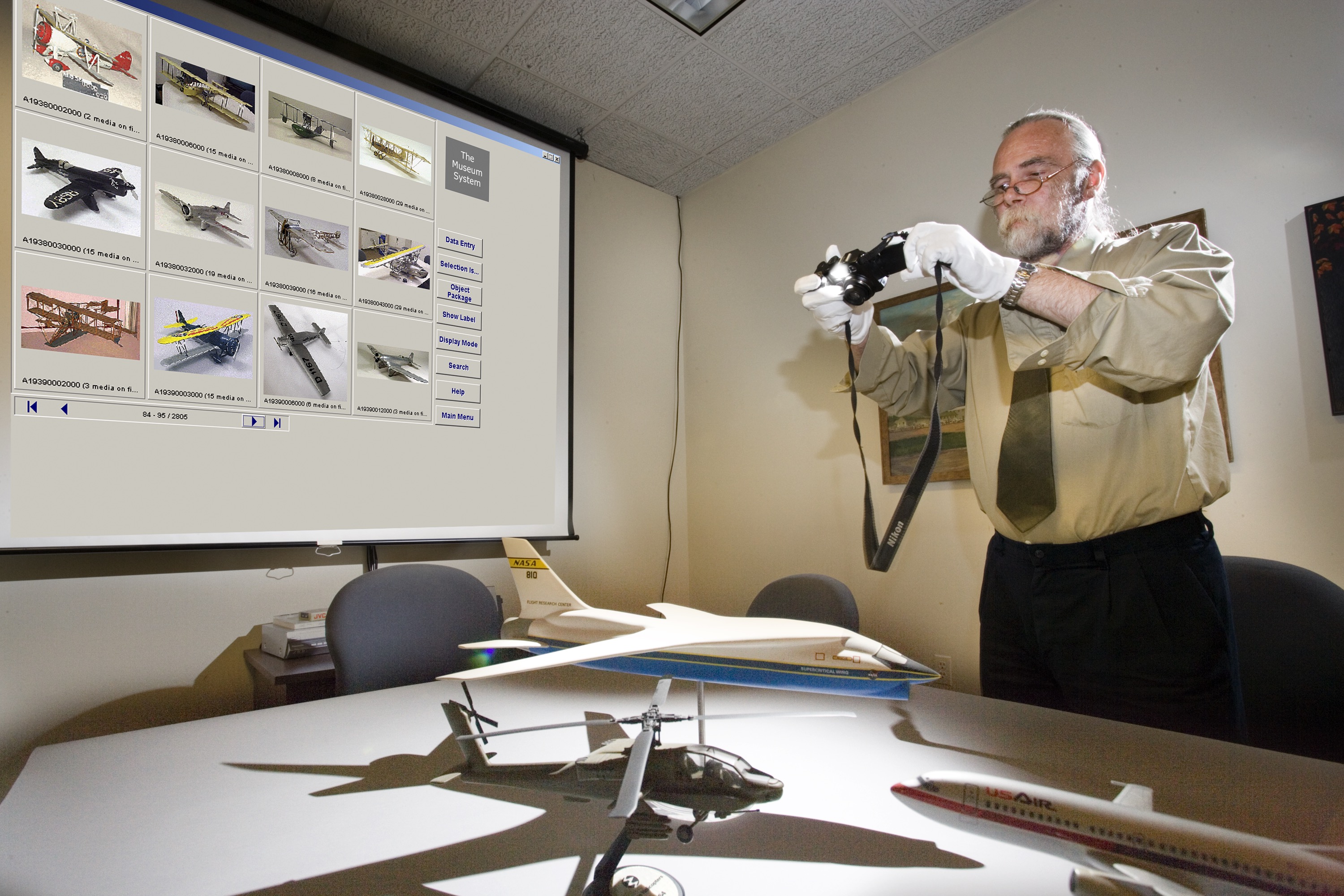 A man stands over a model airplane. He is wearing white gloves and is holding up a camera. 