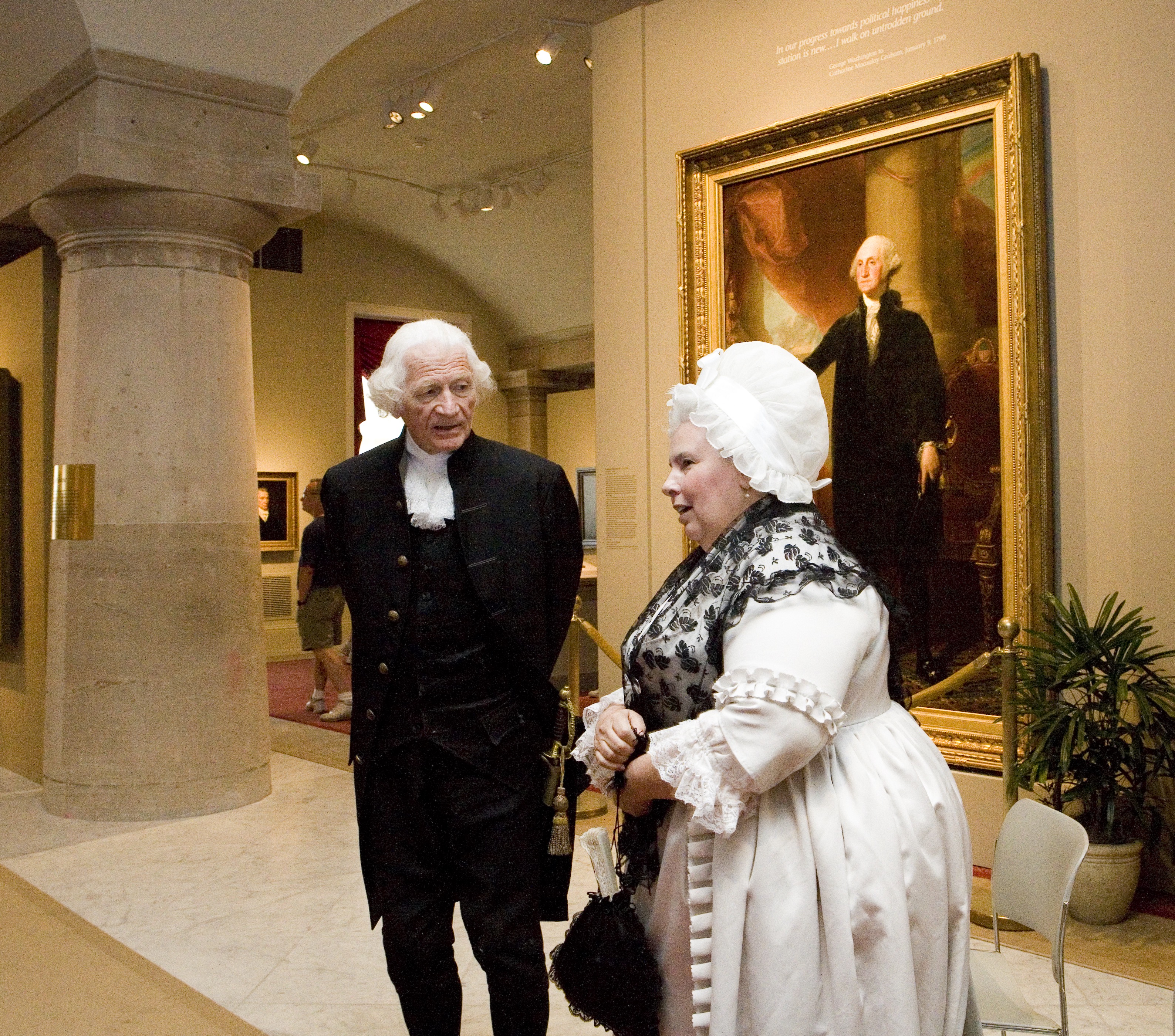 George and Martha Washington impersonators stand in front of a large portrait of George Washington. 