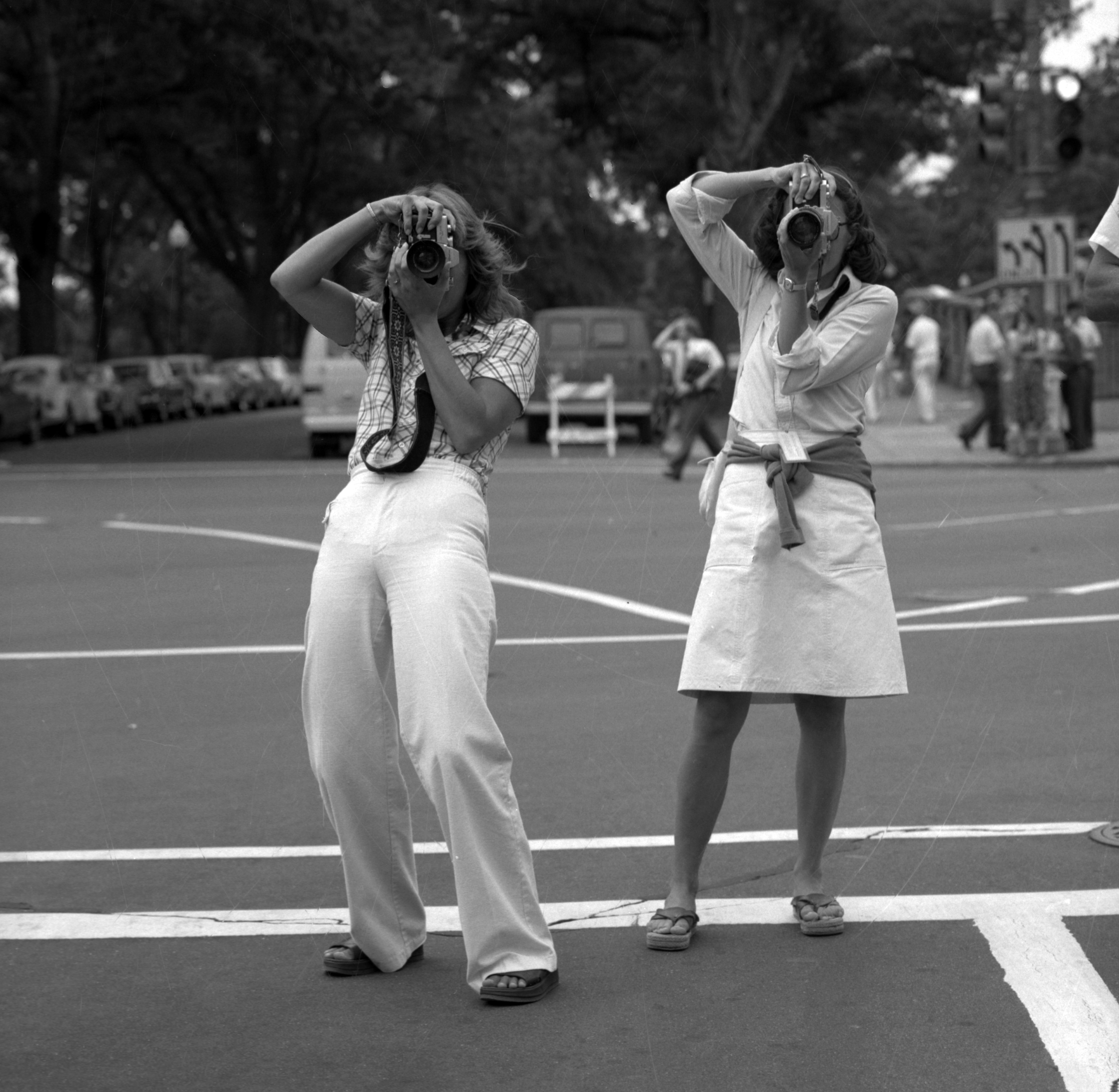 Two women, wearing white, hold cameras up to their eyes. Their faces are not visible. The camera is 