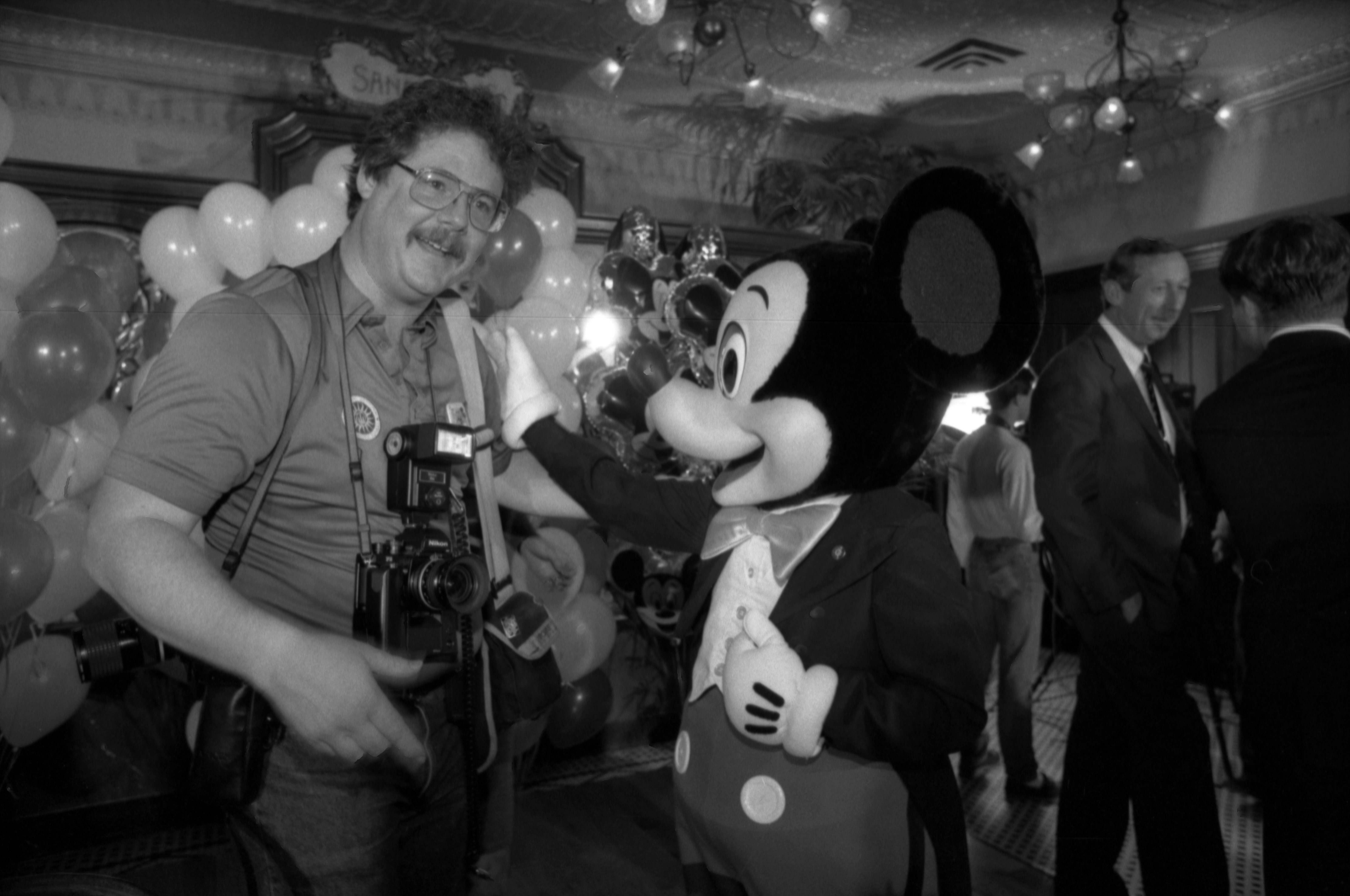 Someone in a Mickey Mouse costume puts a hand on a man's shoulder. The man is holding up a camera. 