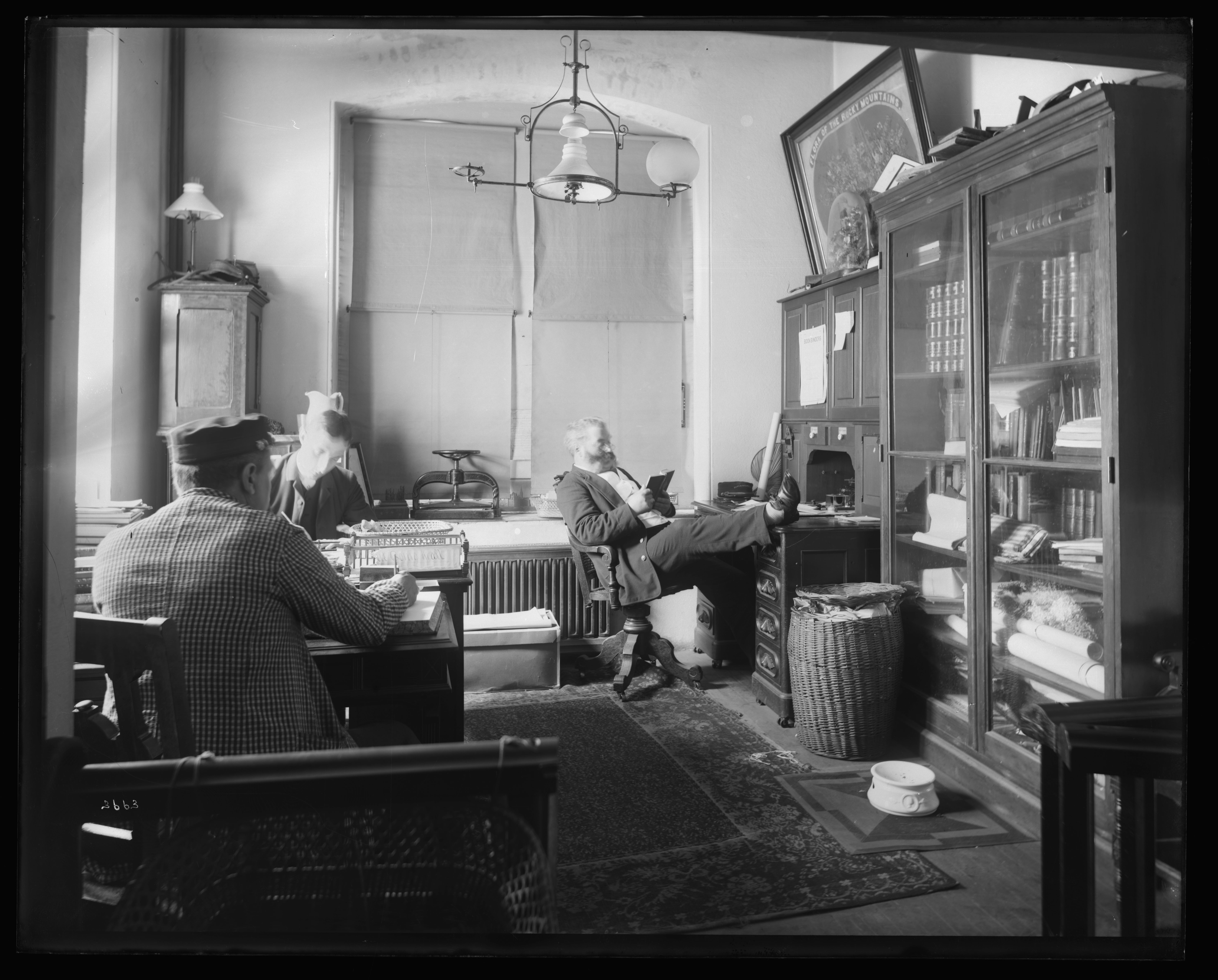 Black and white image of three men sitting at desks in an office space.