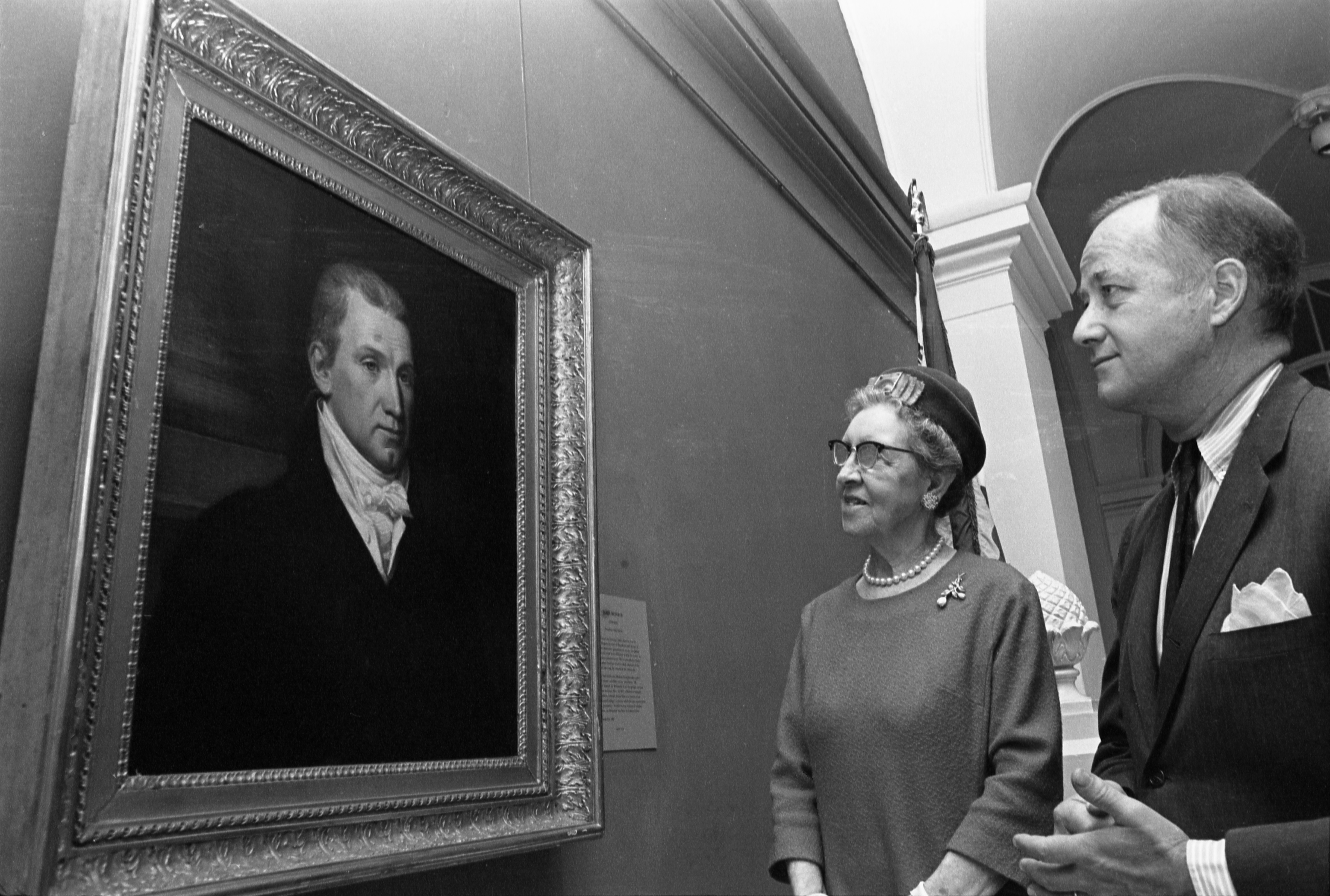 Two people stand in front of a portrait of James Monroe. 