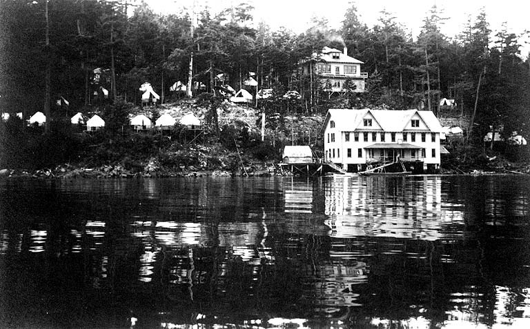 Black-and-white photograph of a building on the water. Another large building is slightly more inlan