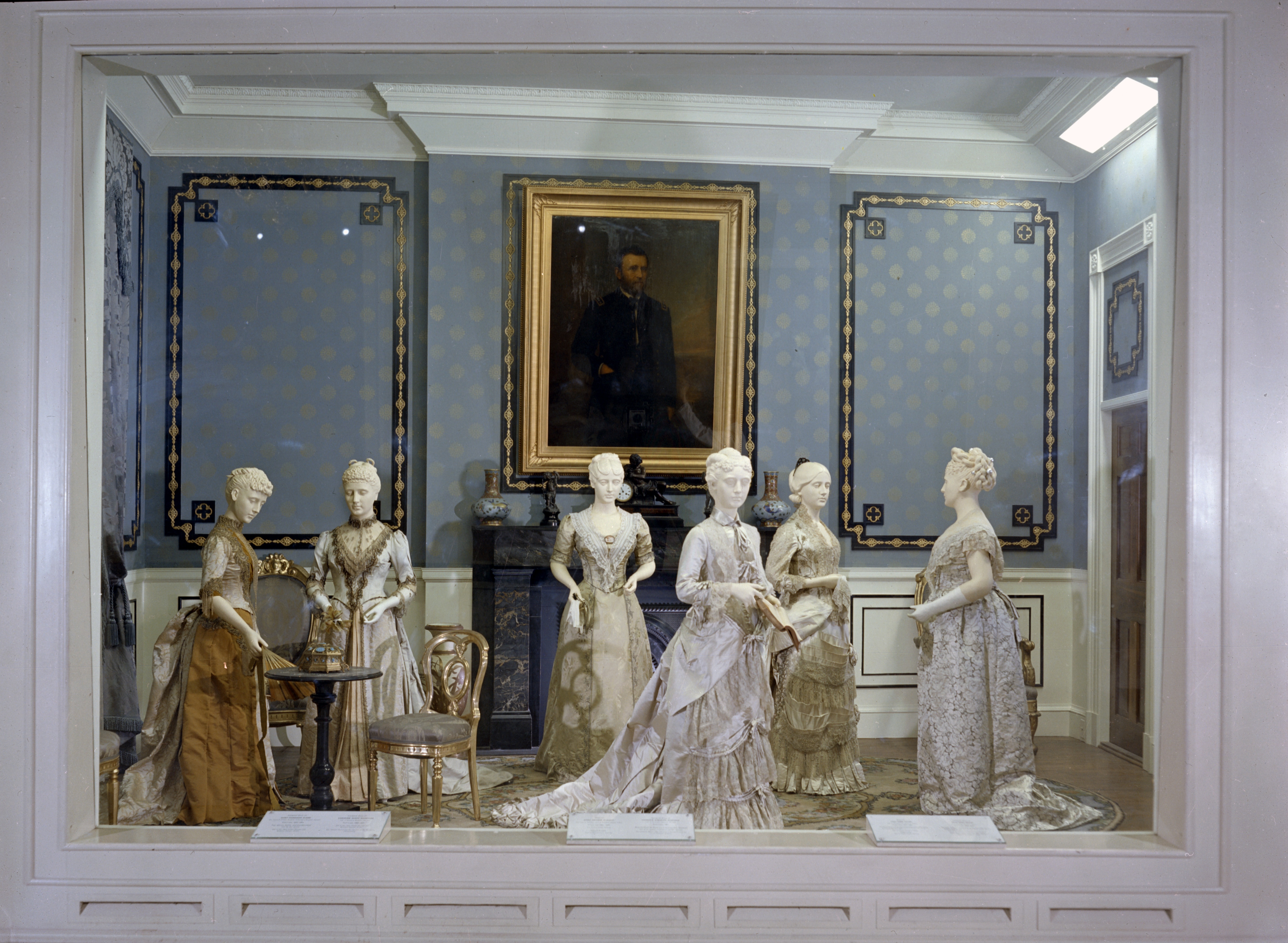 6 mannequins in gowns stand in a blue room with a portrait in the background on a wall. 