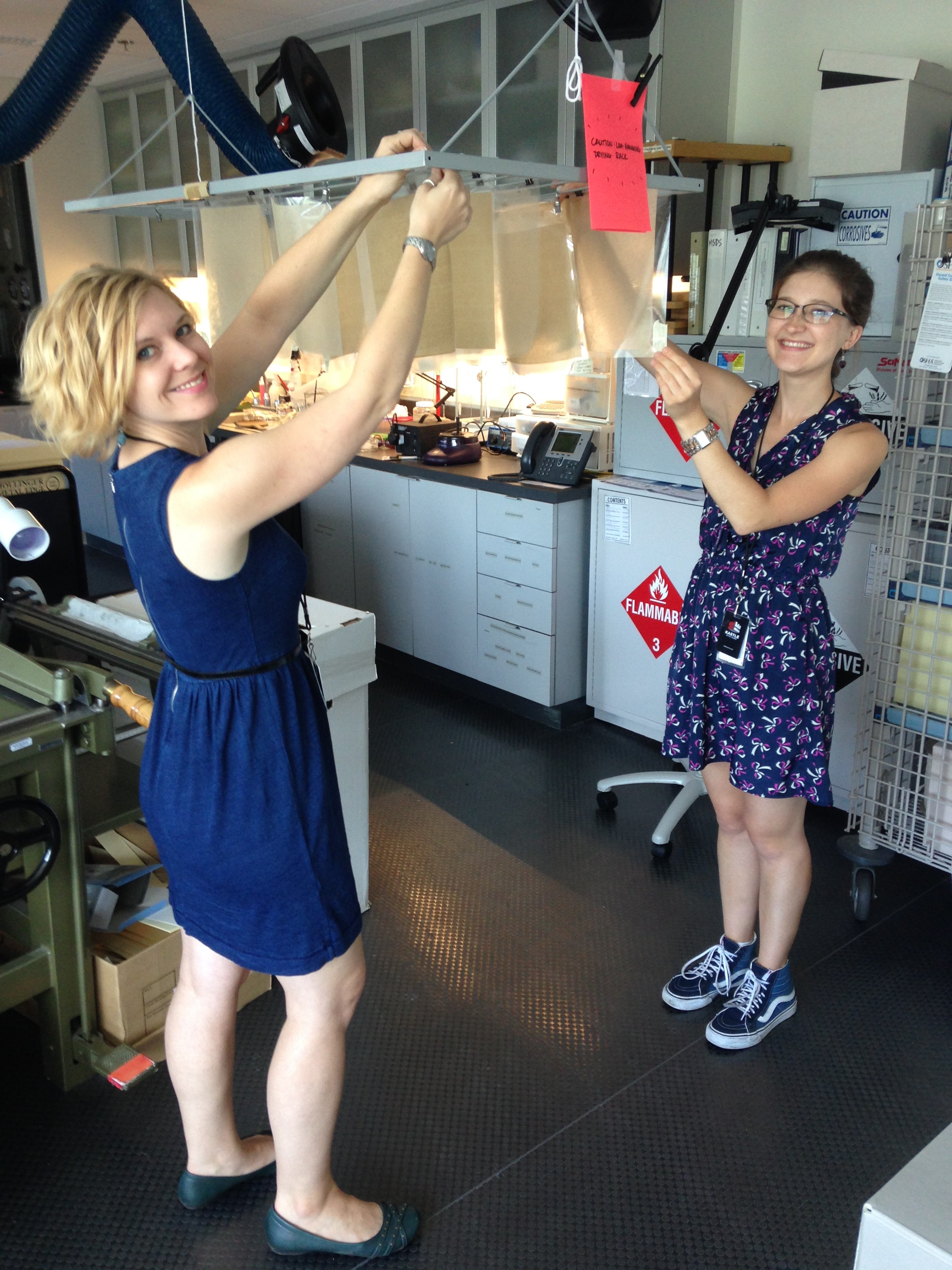 Two people stand in a lab and hang pieces of paper vertically, 