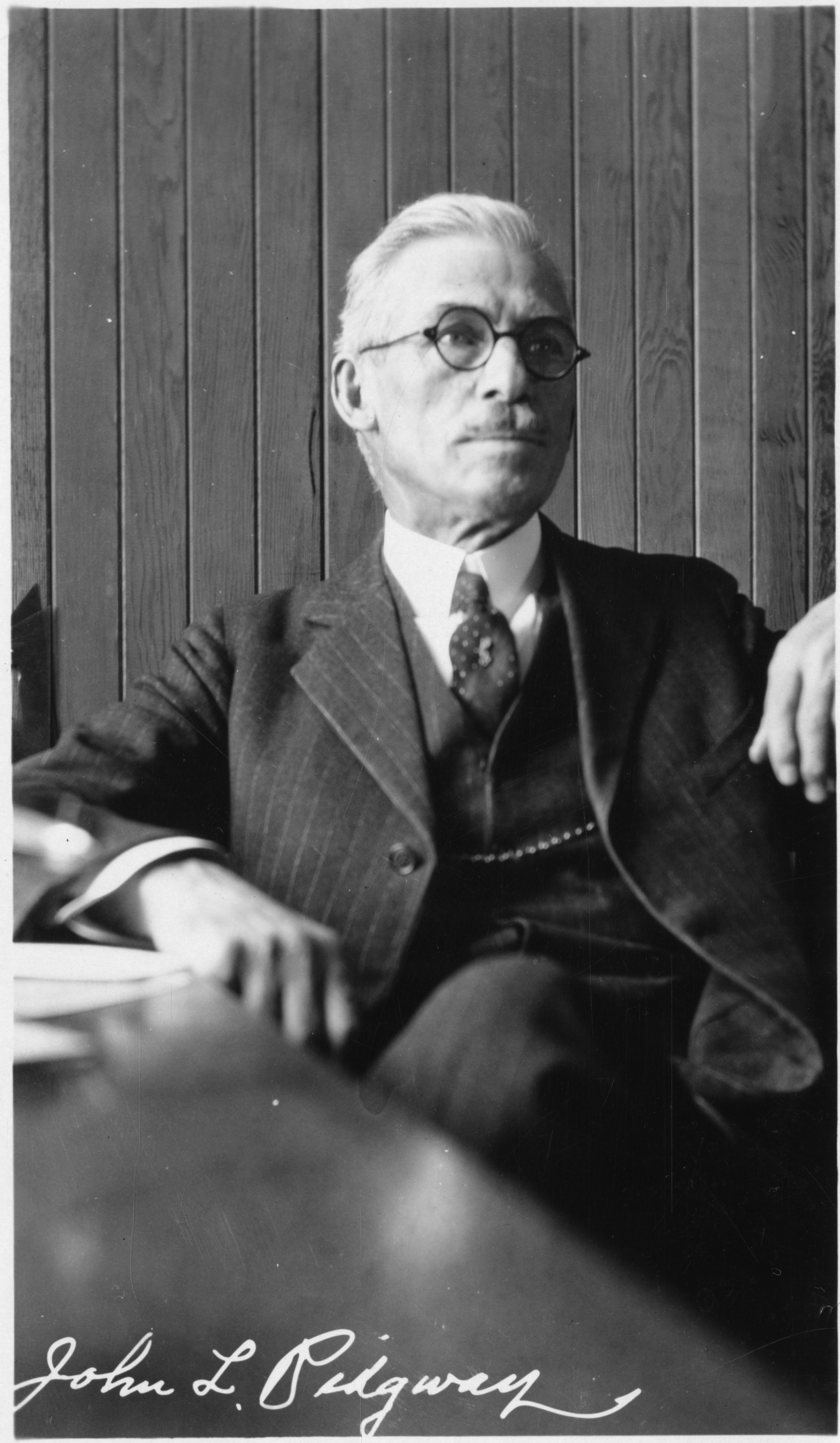 A man in a suit and wearing glasses sits at a desk. He leans back. At the bottom of the photograph "