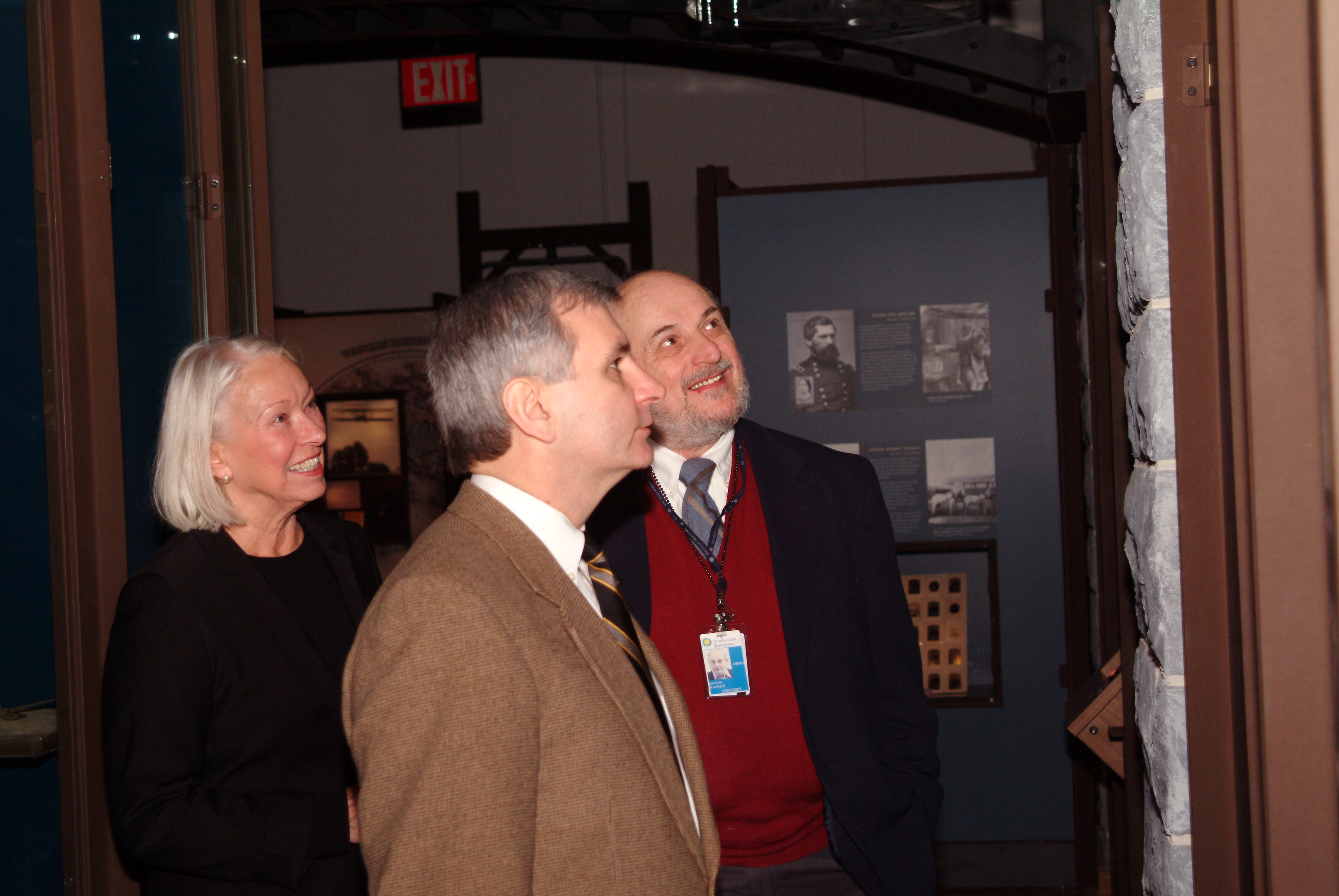 One woman and two men view a wall in an exhibit at a museum. The wall appears to be made of stone.