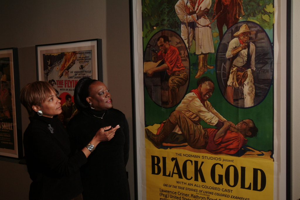 Two women stand near a poster for Black Gold. The woman on the left is pointing toward the poster.