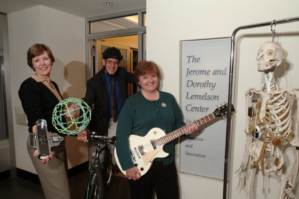 Three people pose with technology. Klose is holding up a circular toy that folds in and out and a foot scale. The man in the middle is riding a bike and weaning a helmet. The woman to the ride is holding a guitar.