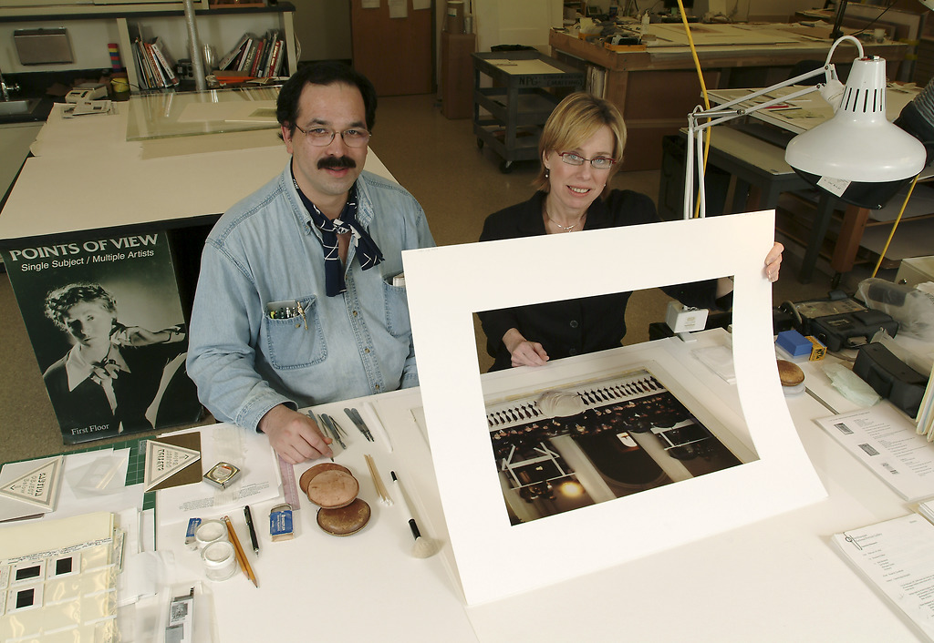 A man sits next to Rosemary Fallon, who is lifting a mat from what appears to be a photograph on a desk. Weights hold down the image. Both people are looking at the camera. 