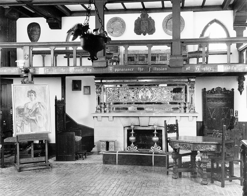 B&W photo of interior with fireplace, a portrait of a woman on an easel, and highly ornate wood furniture.