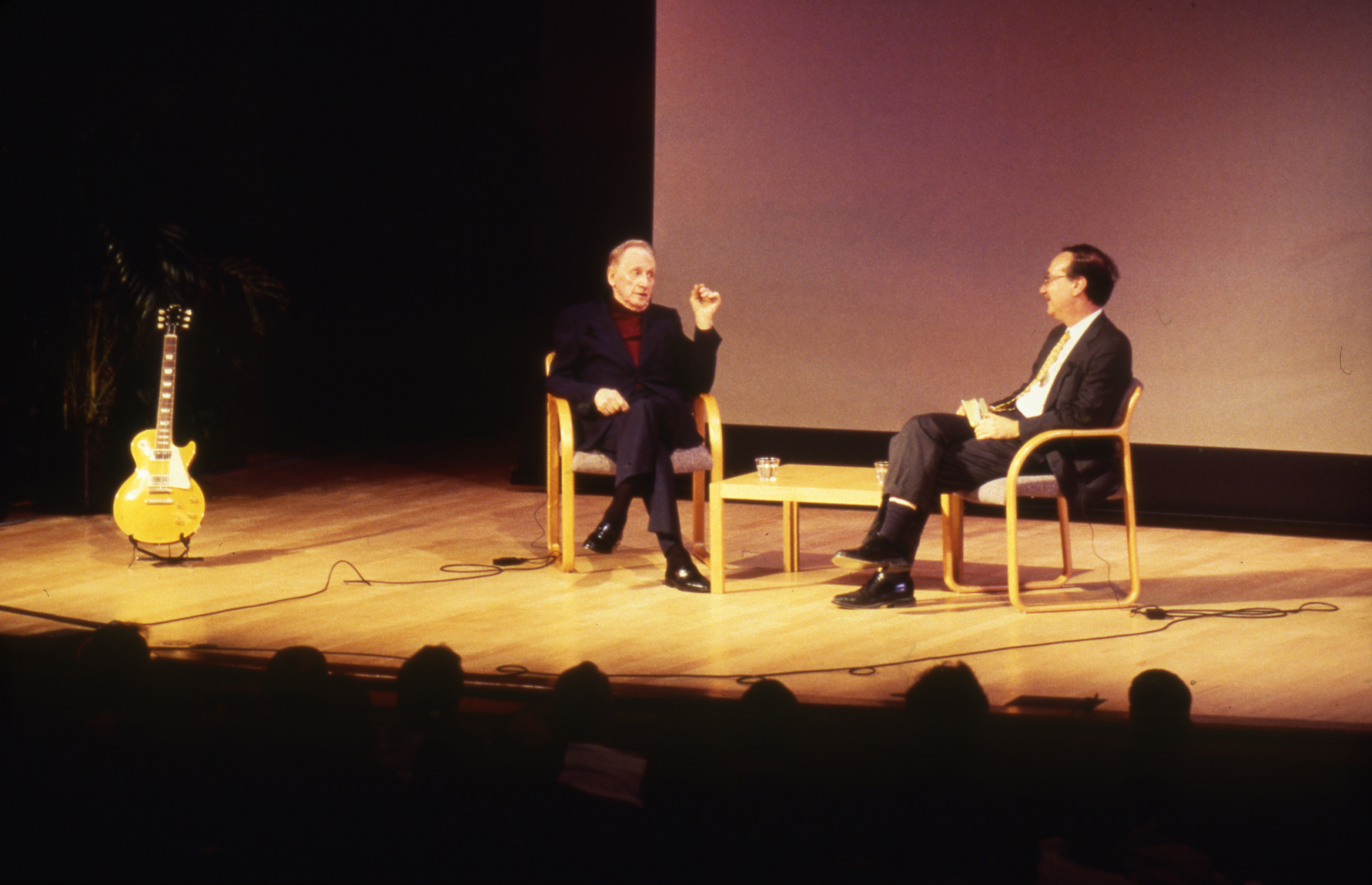 Les Paul and March Pachter seated on a stage having a conversation with a Les Paul guitar in a stand