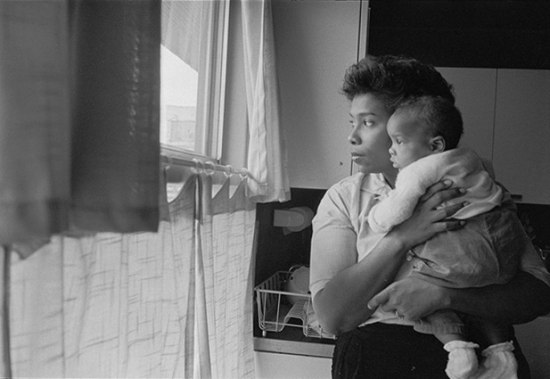 Black and white image of African American woman holding her baby, looking out the window.