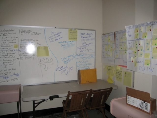 Whiteboards and poster boards are covered in post-its. It’s difficult to make out what they say. Temporary desks are below the boards.