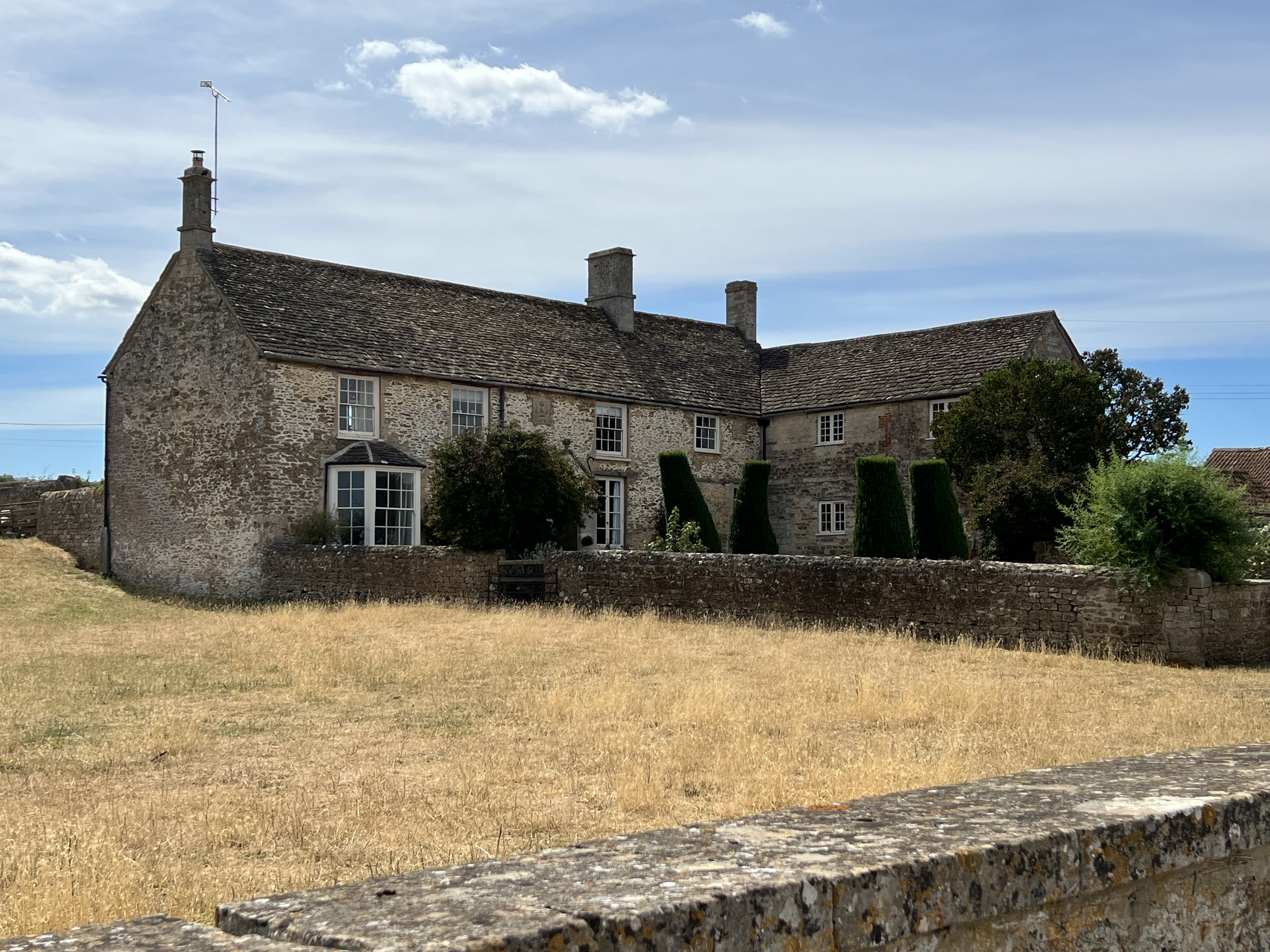 An L-shaped stacked stone and mortar house with a slate roof and white-painted sash windows sits ami