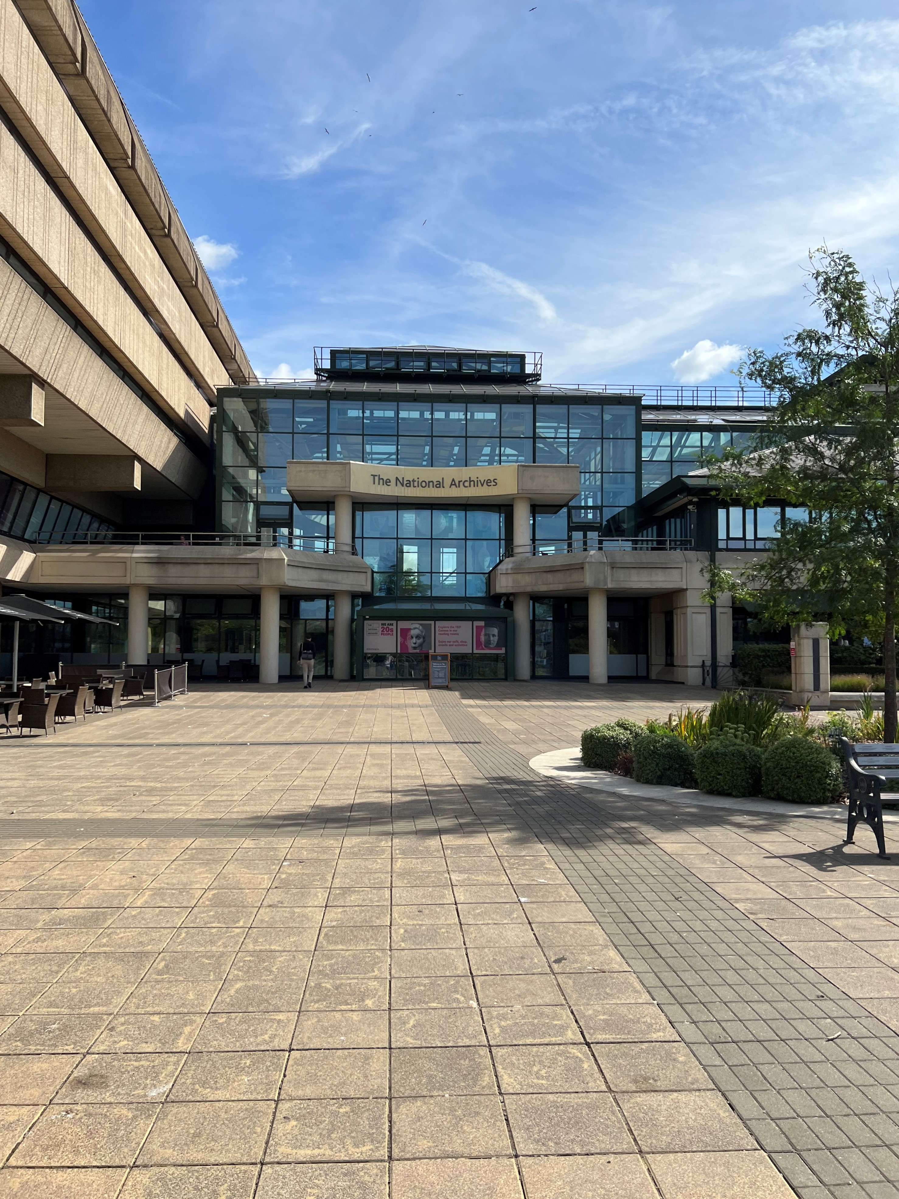 A modern glass atrium sits adjacent to a bulky concrete building, fronted by an expansive paved cour