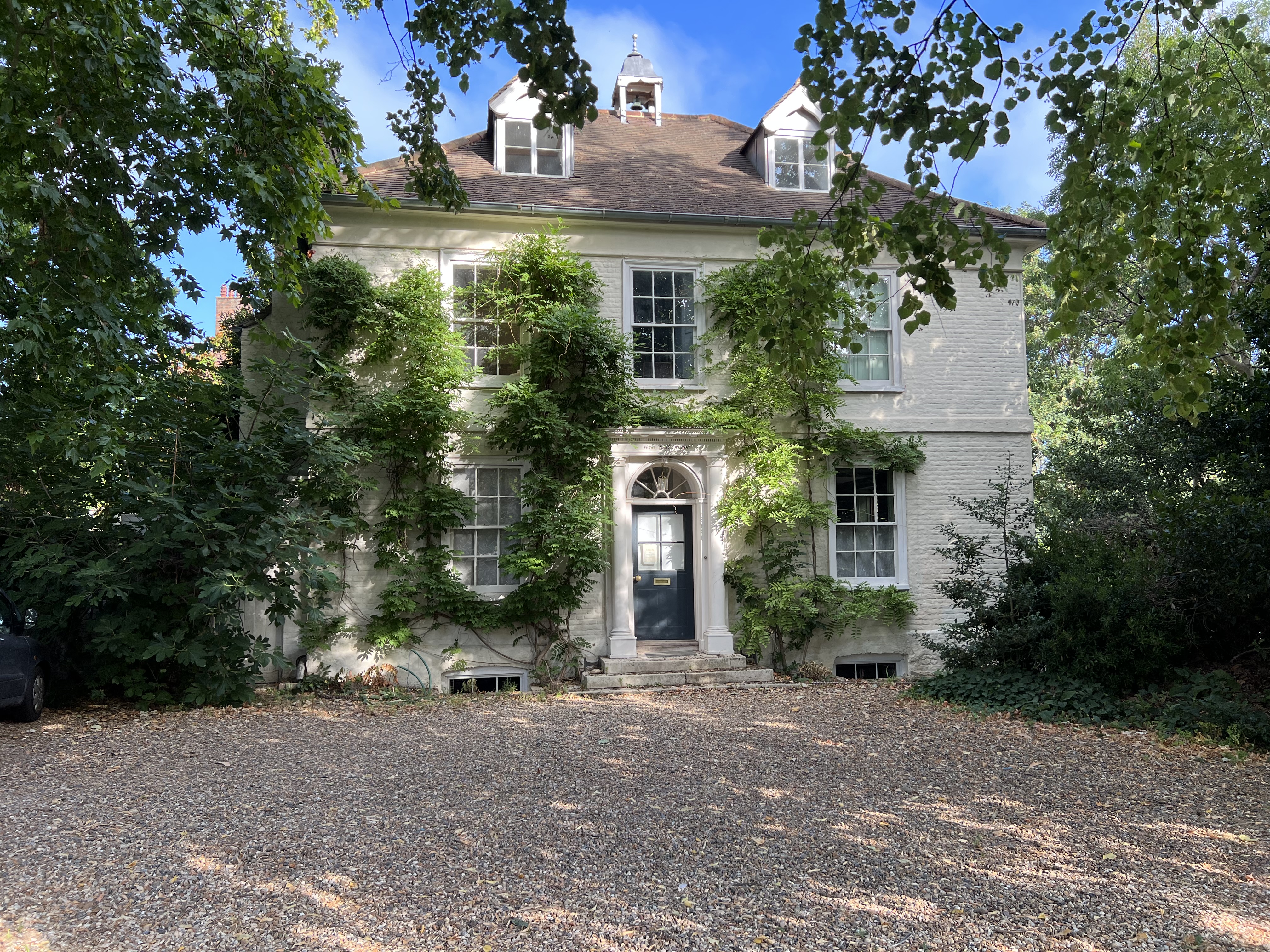 A two-story white brick home with tile roof and twin dormers is slightly overgrown with climbing vin