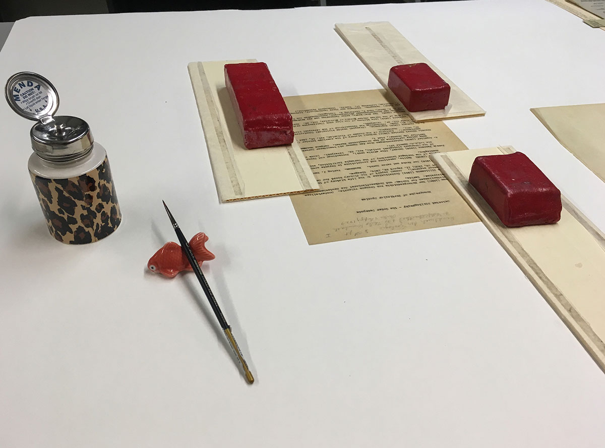 Archival document held by weights with artist brush nearby.