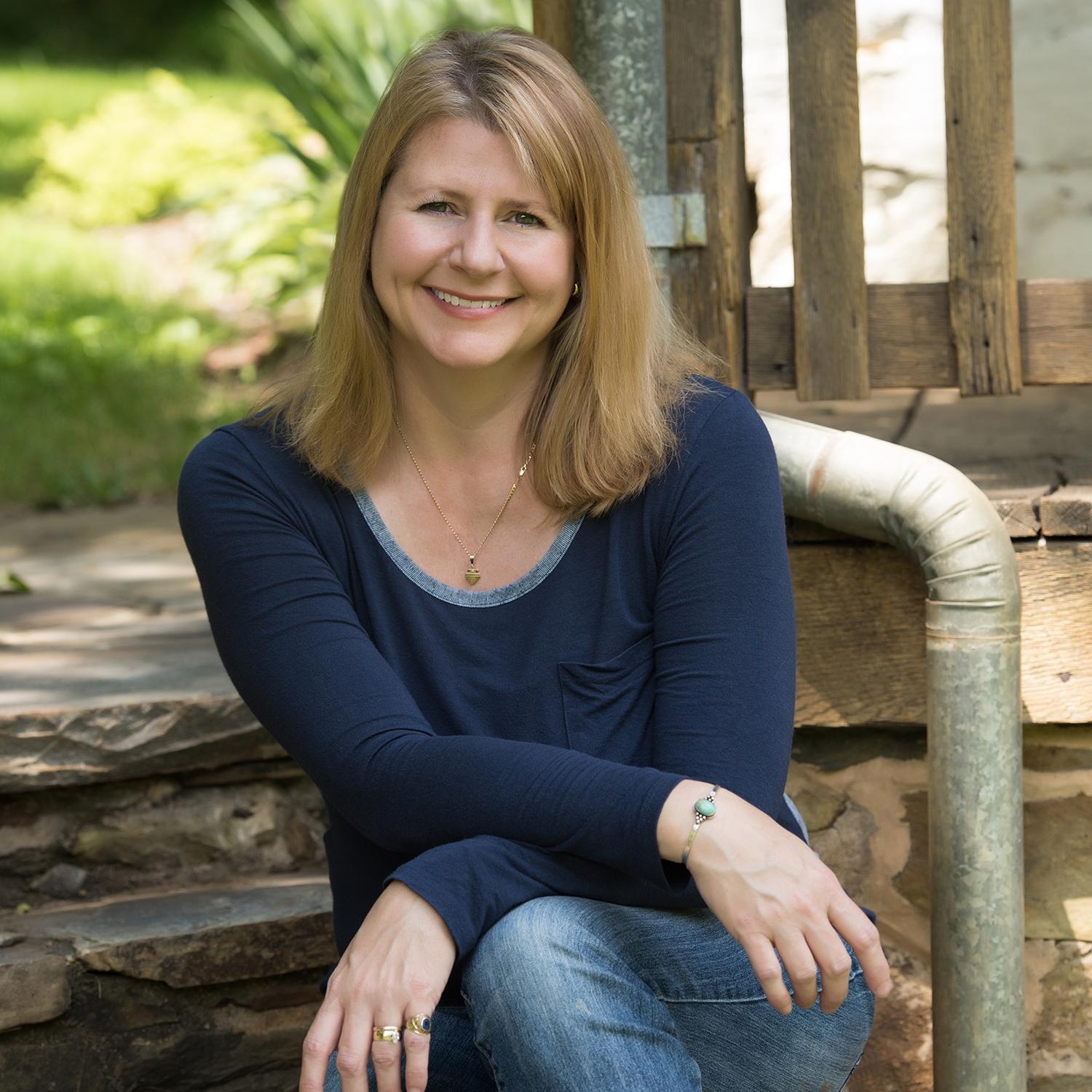 A white woman in a blue shirt and blue jeans wearing rings, necklace, and bracelet sits outside on a