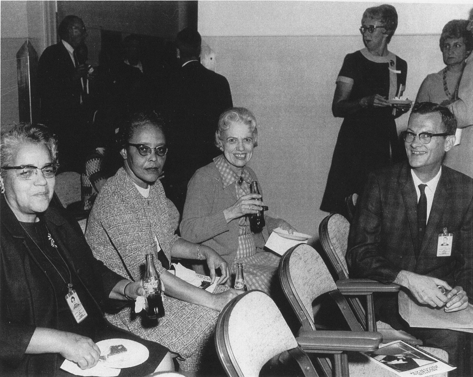 Black and white photo of 3 seated women and 1 man at reception holding plates and drinking Coca Cola.