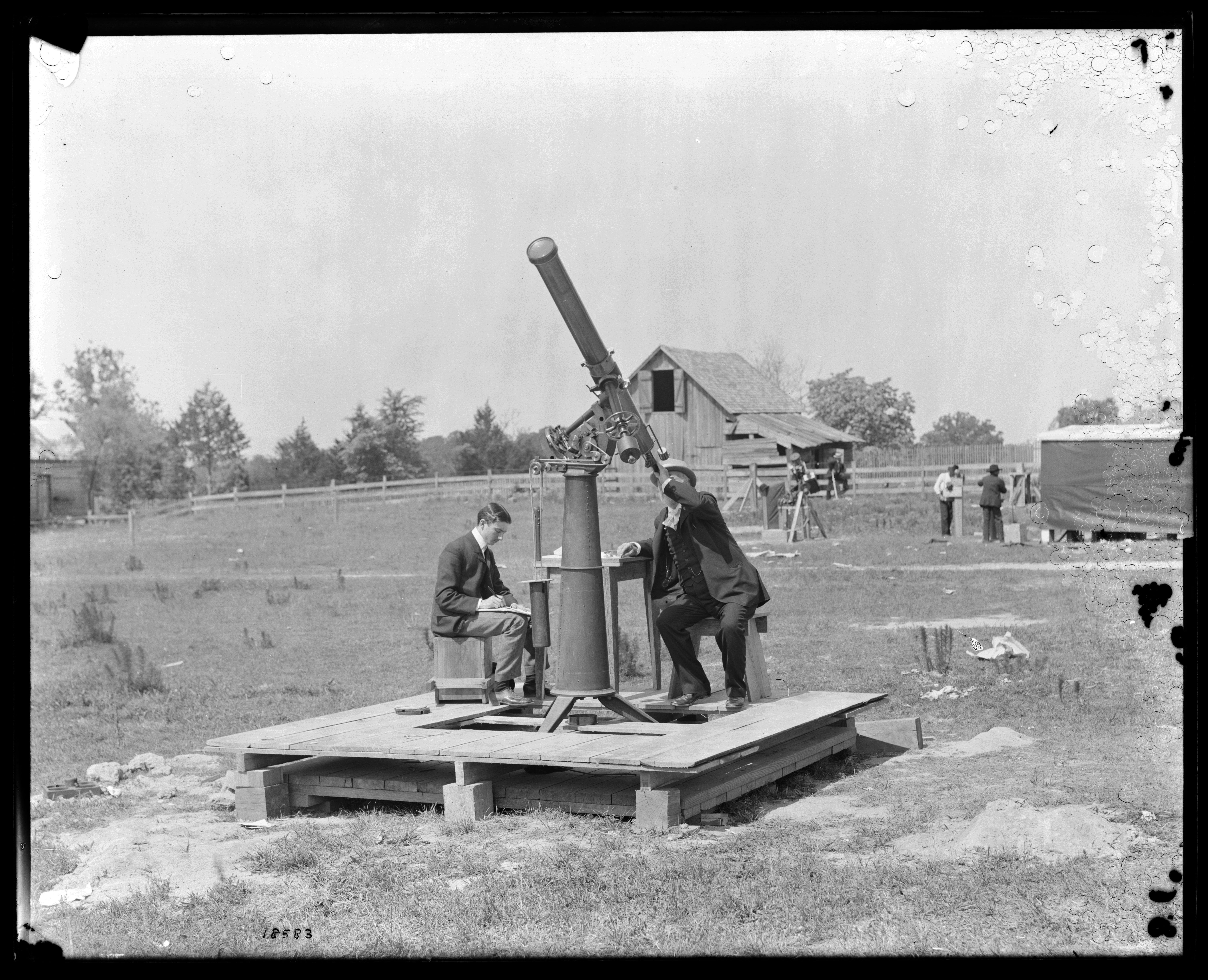 2 men in a field on wooden platform with telescope with one man peering up into the telescope.