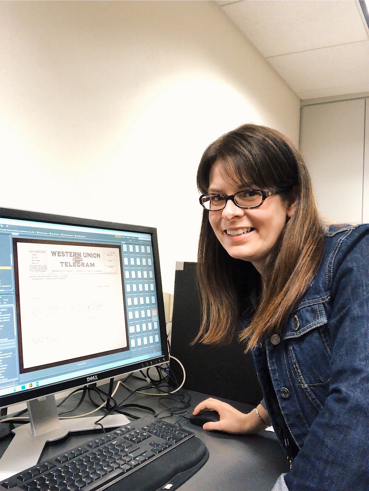 A woman wearing glasses and a denim jacket posed for a picture next to a computer screen. She has on hand on a computer mouse.