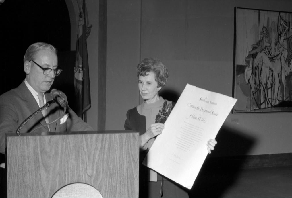 Man in front of podium talking with woman beside him holding large certificate