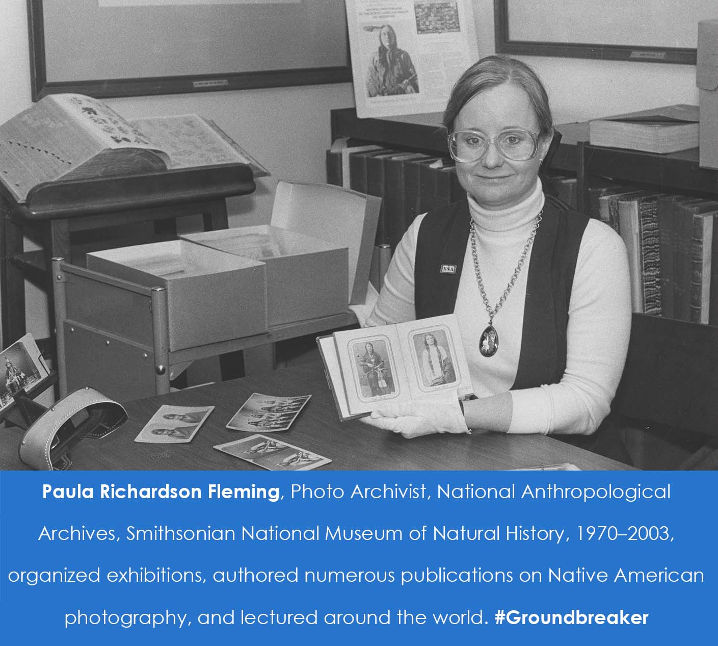 A woman sits at a desk and holds up photographs of Native Americans. She is wearing white gloves. Archival boxes are in the background.