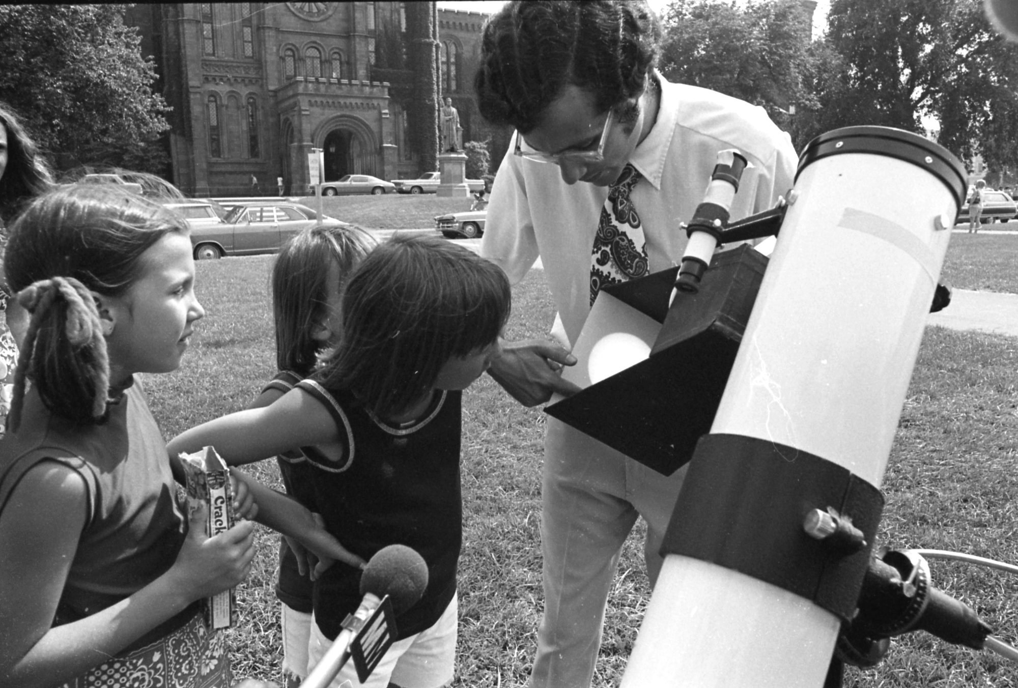 B&W photo of man demonstrating telescope with 2 children observing.