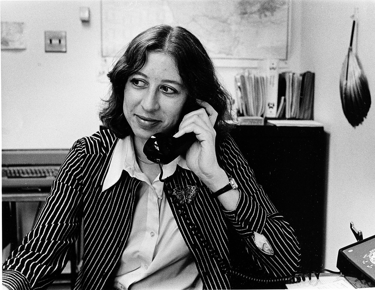 A woman is seated at a desk and holds a telephone to her ear. She is wearing a striped jacket. She is smiling though looking off to the side, not exactly toward the camera. 