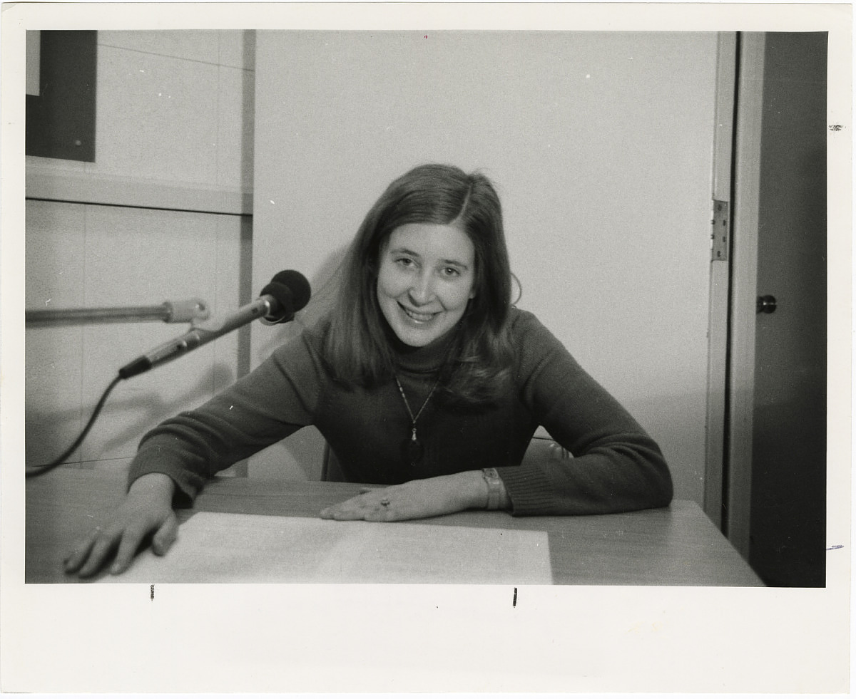 A portrait of a woman seated a desk. She is smiling toward the camera. A microphone hangs near her h