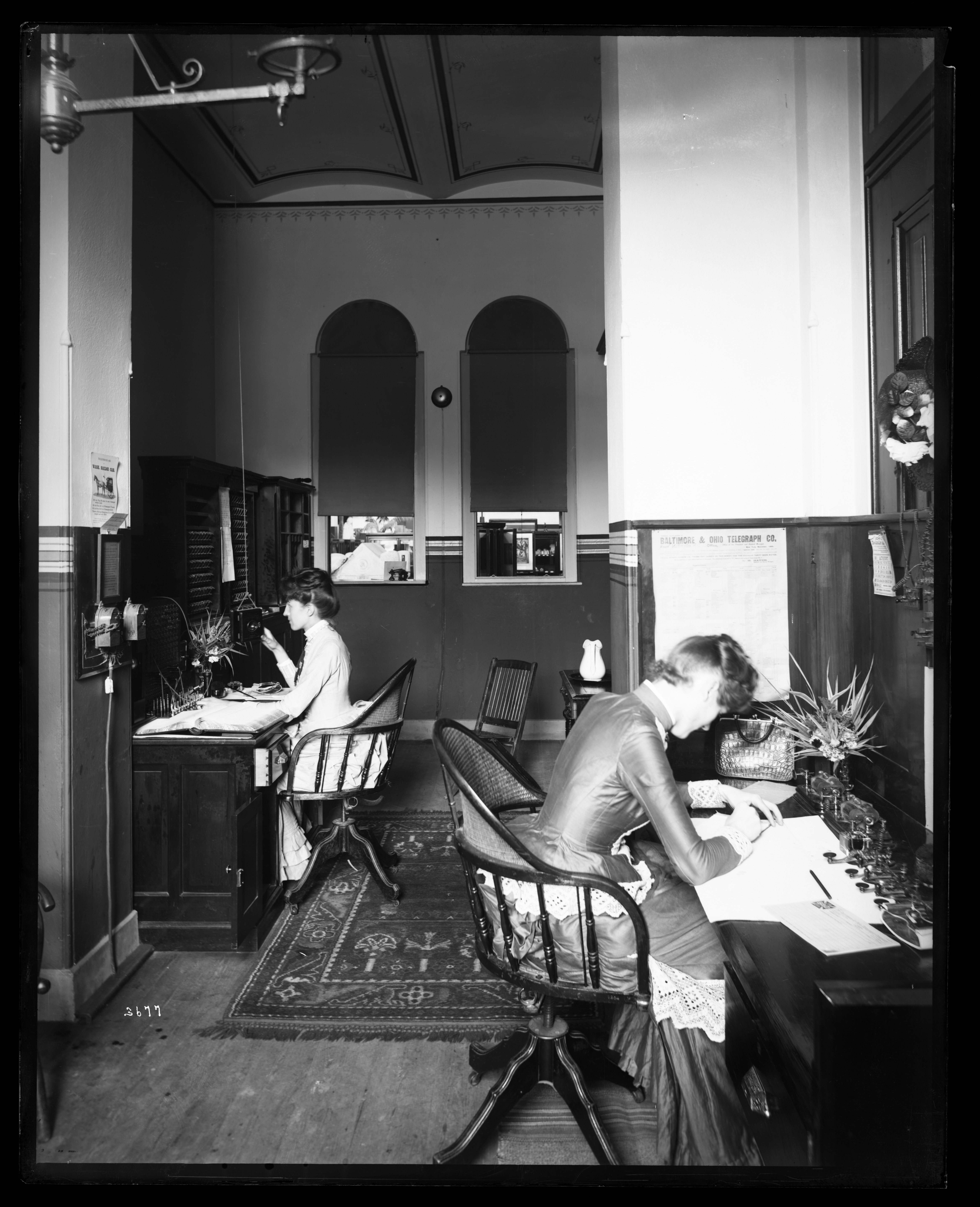 Two woman in long dresses sit and work at desks. 