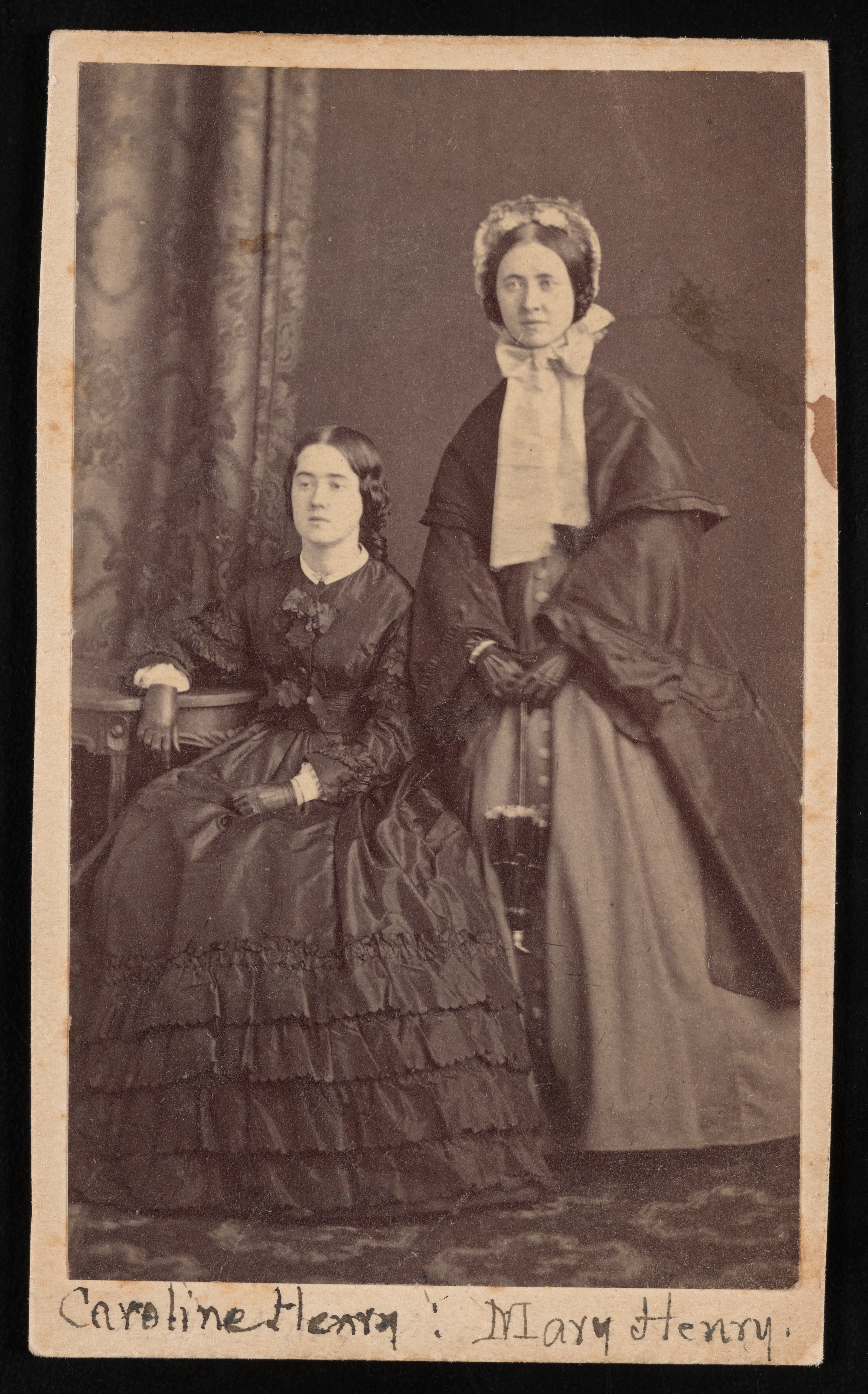 Caroline Henry is seated and Mary Henry stands behind her. Both women are wearing long dresses and not looking directly at the camera. 