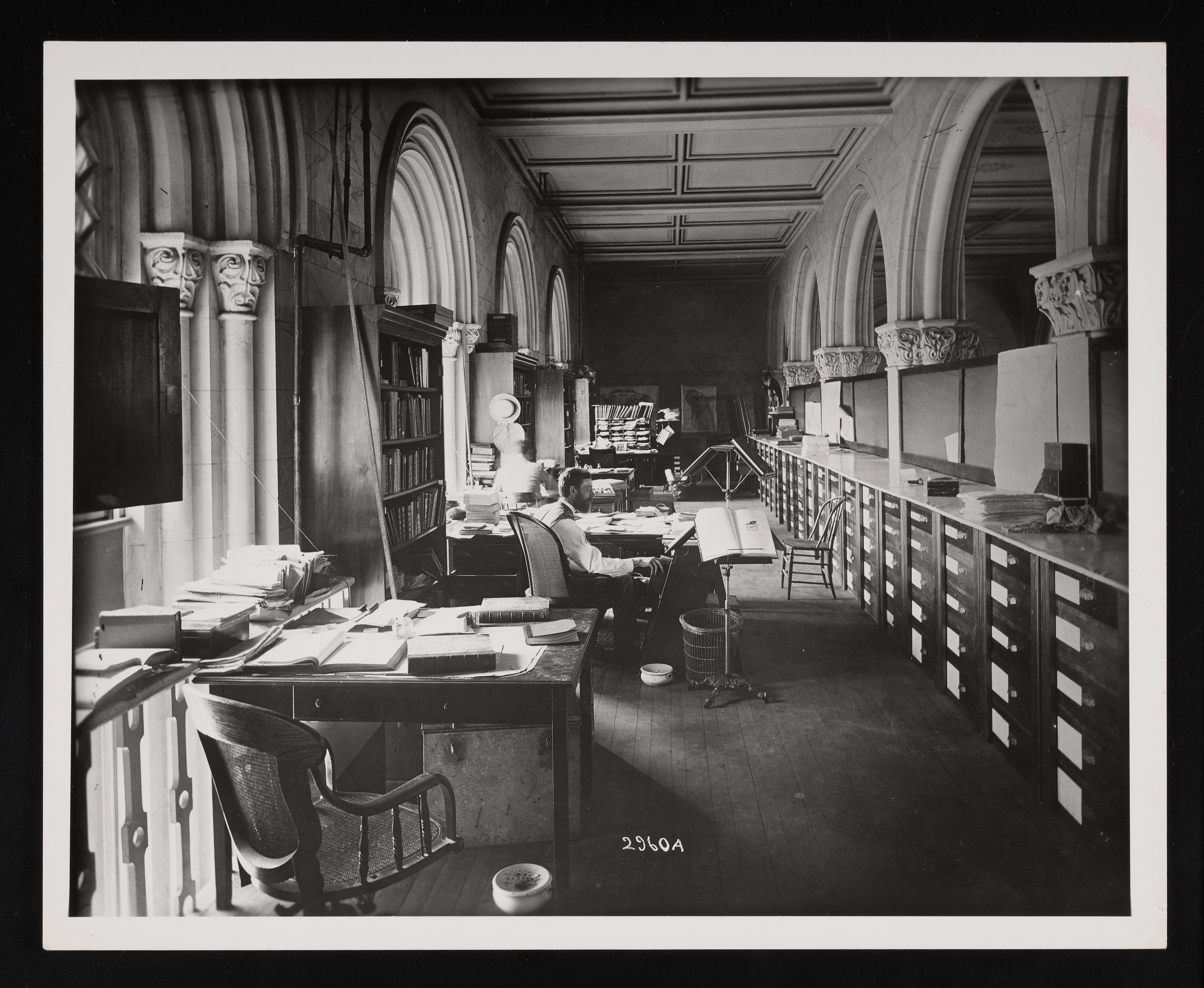 Office space with one man working at the second desk closest to the camera. Books are on the desks. There is collections storage on the right side.