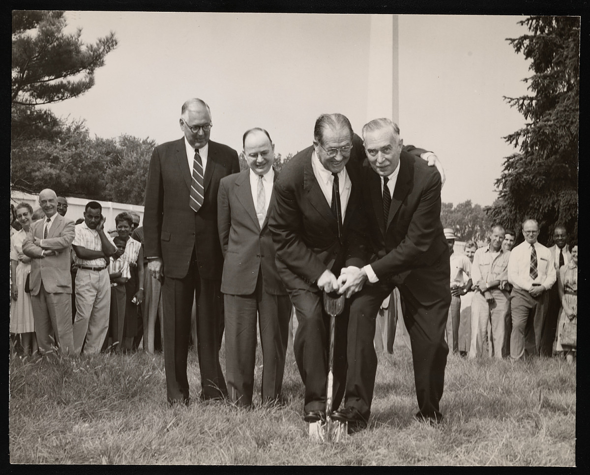 Four men stand near a shovel. Carmichael is holding the shovel with both hands. Another man has one 