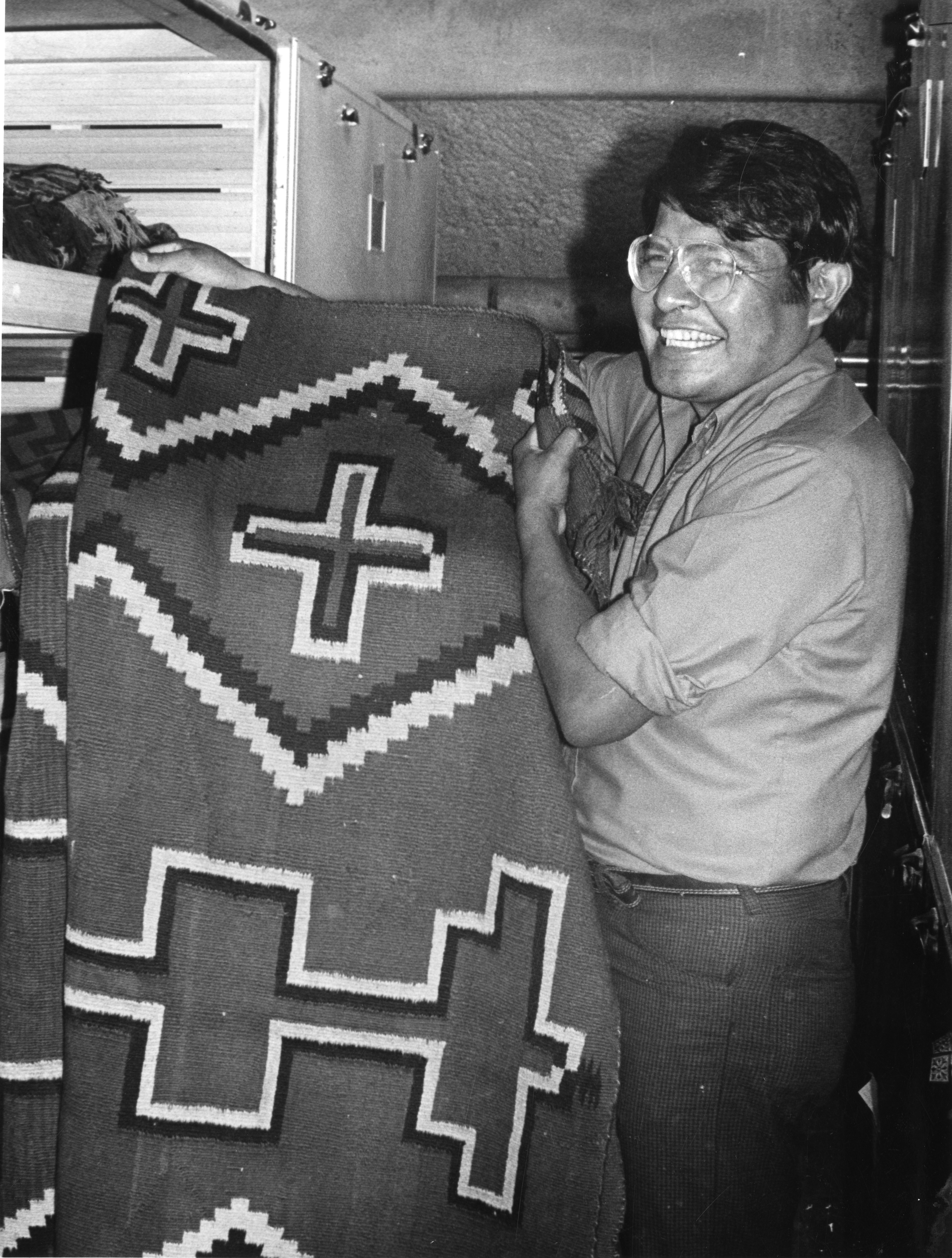Black and white image of Harry Walters, Navajo male, holding a blanket in a museum collection storage area.