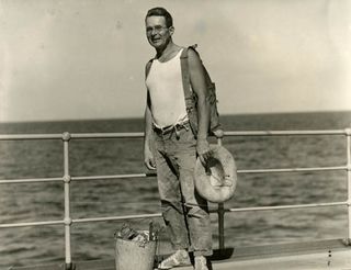 Black and white image of man standing on boat with ocean in the background.