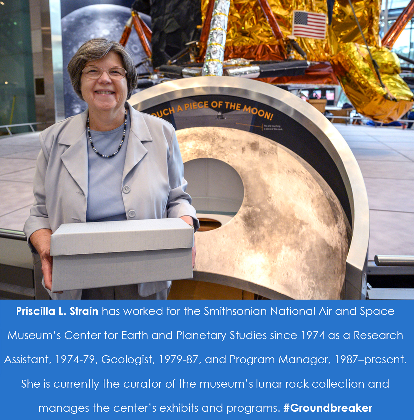 A woman holds a box and smiles at a camera. She is standing in front of a lunar rock sample.