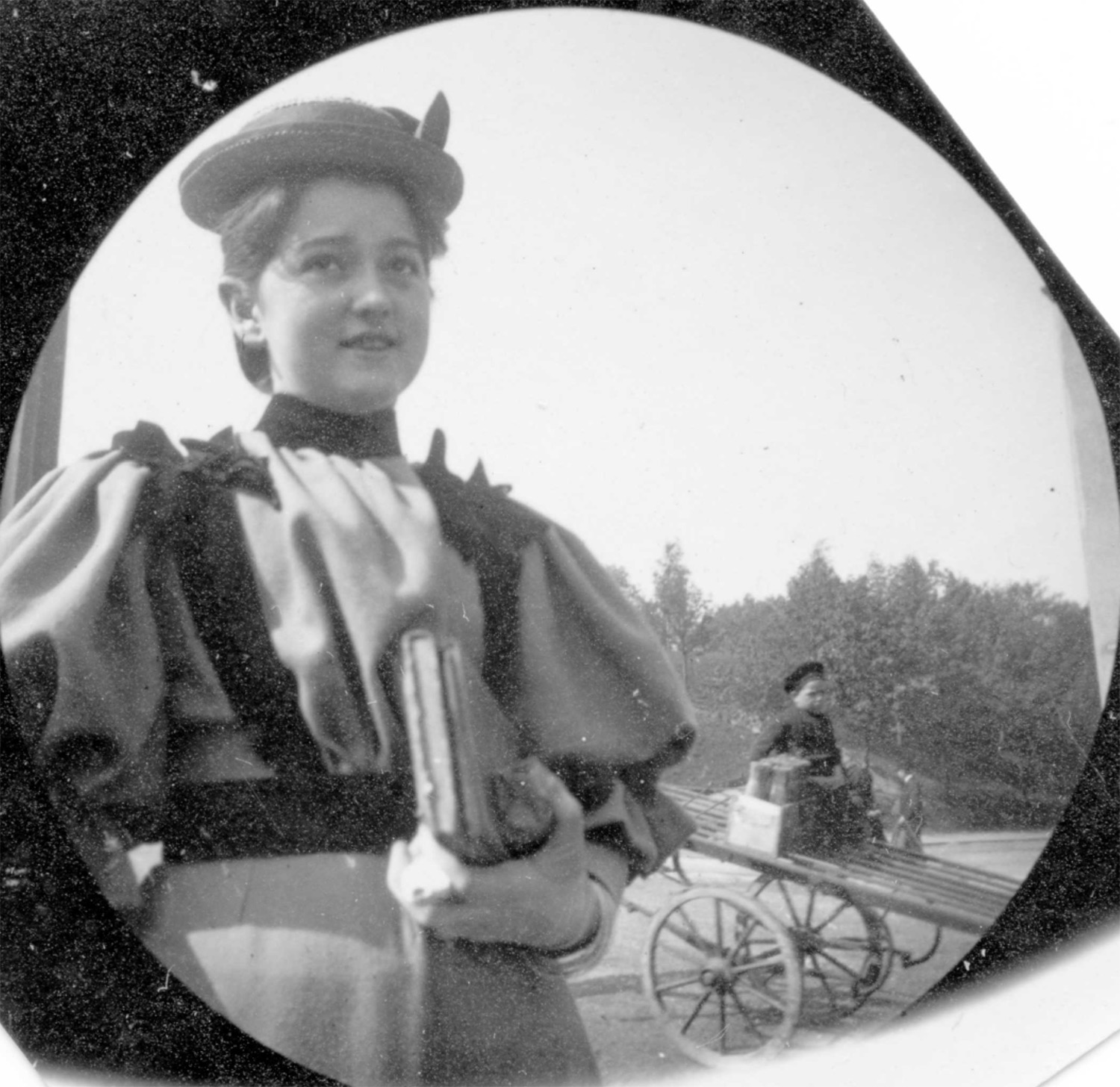 Girl in fancy hat & long dress carrying book with boy on a tipped cart in the background.