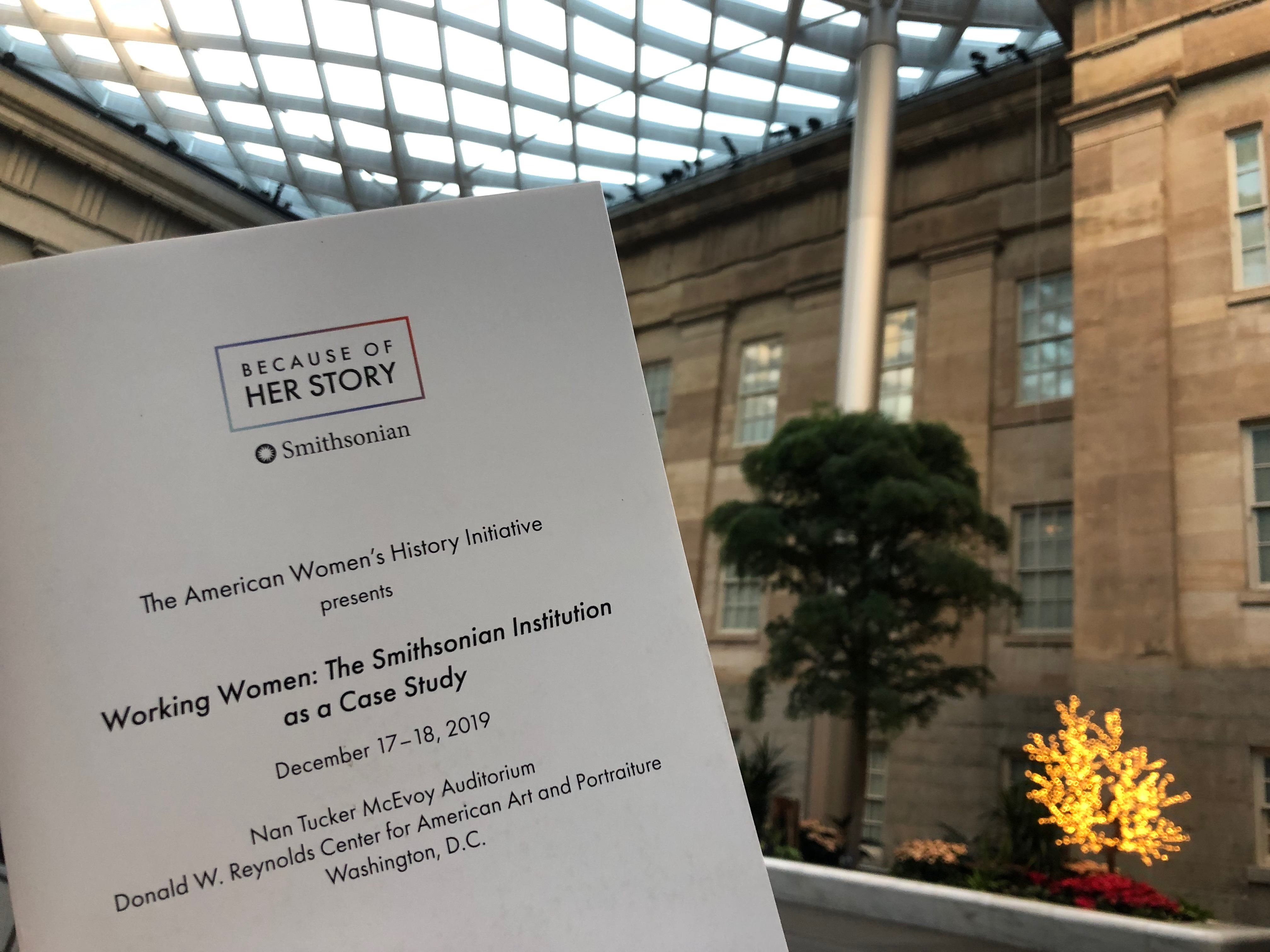 Someone holds up the program for the symposium. The ceiling at the Kogod Courtyard is visible in the background.