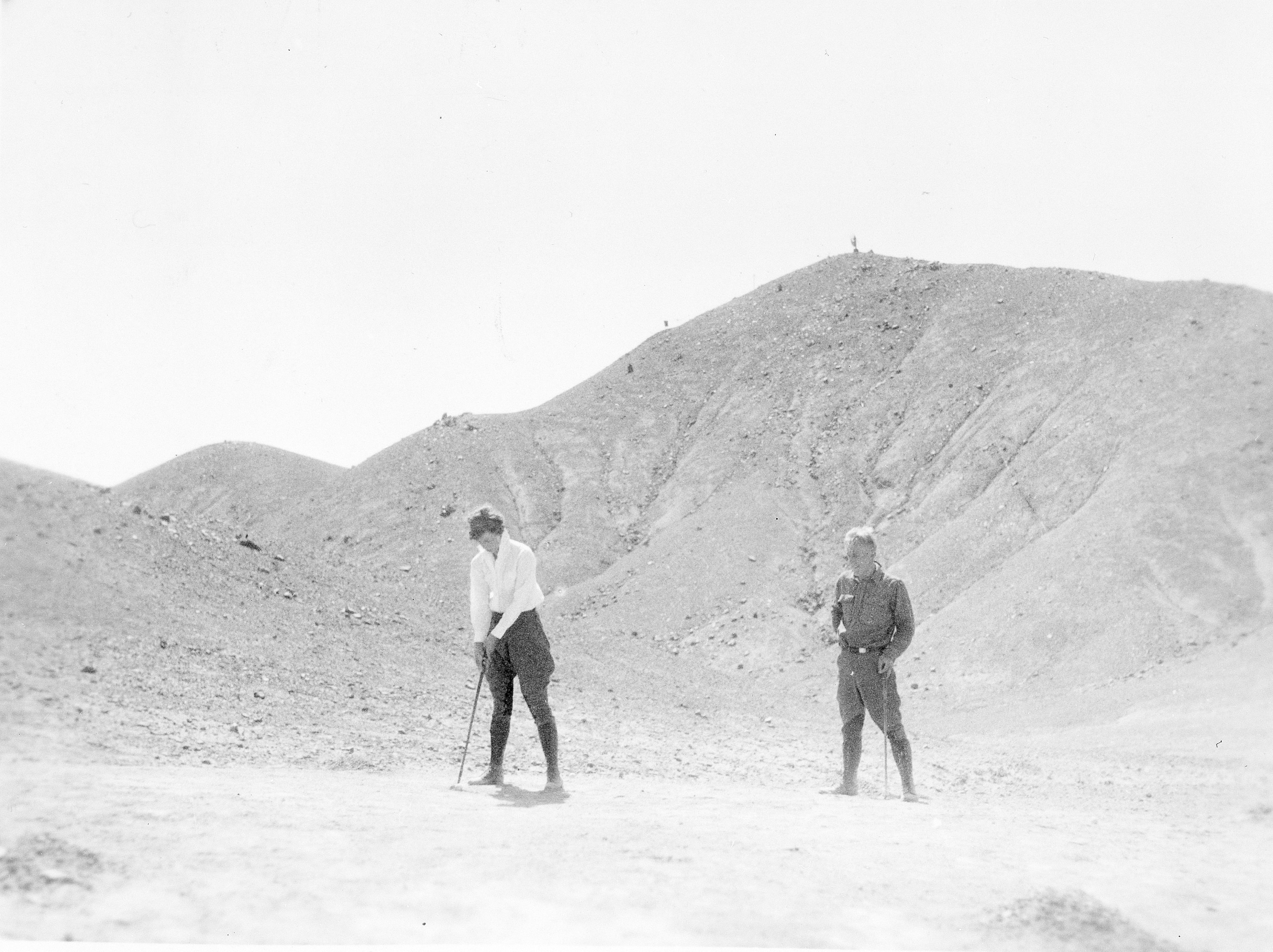 A man and woman stands outdoors in a rocky area. Both are holding the golf clubs and the woman is preparing to take a swing.