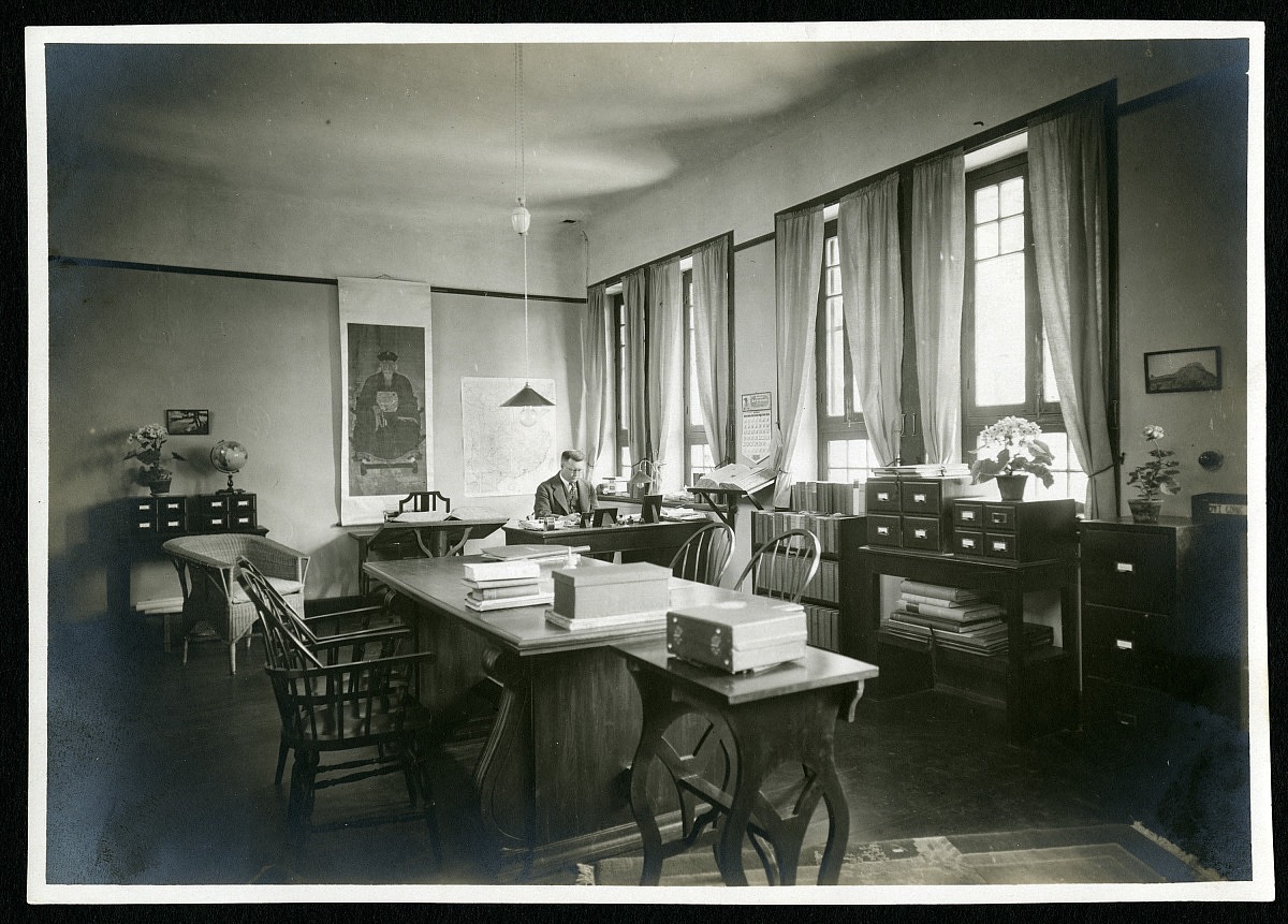A man sits at a desk in an office. Papers and books are stacked on desks in the office. 