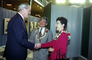 Color image of three people indoors, two of which are wearing leis and one is shaking the hand of an