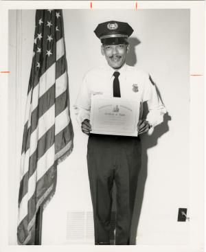 A guard in uniform smiles at the camera and holds up a certificate. He is standing next to an Americ