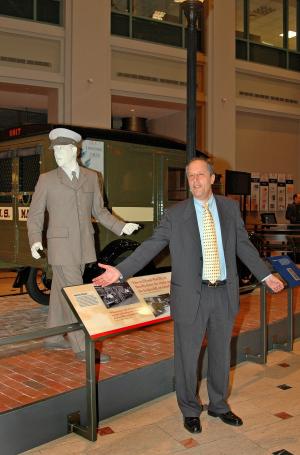 Man in a gray suit stands in from of a mannequin dressed as a postal worked and postal vehicle.