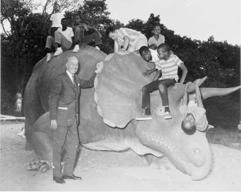 Black and white photograph of man standing next to sculpture of Triceratops with several young child