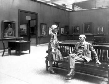 Black and white photograph of a man sitting on a bench and a women leaning one leg on the bench in the middle of a room filled with portraits at a museum. 